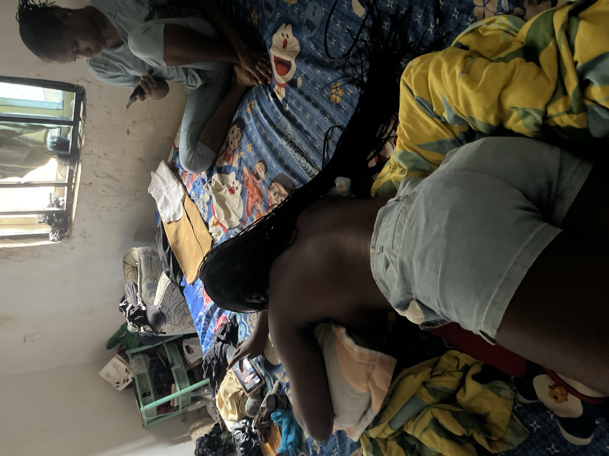In Takau, Nigeria, a room exhibits a state of disarray. Two young women occupy the space, both engrossed in their mobile phones. One woman is seated on a bed, wearing light blue pajamas, her attention fixed on the device in her hands. The second woman is lying on her stomach on the same bed, facing away from the viewer, also looking at a phone. She is wearing light blue denim shorts and her dark hair is braided, extending down her back. The bed is covered with a blue sheet featuring cartoon characters. Clutter surrounds the bed, with various items of clothing and personal belongings scattered across the floor and on a green plastic shelf unit. The room has a window with bars, allowing natural light to enter, and the walls appear to be unfinished or aged.