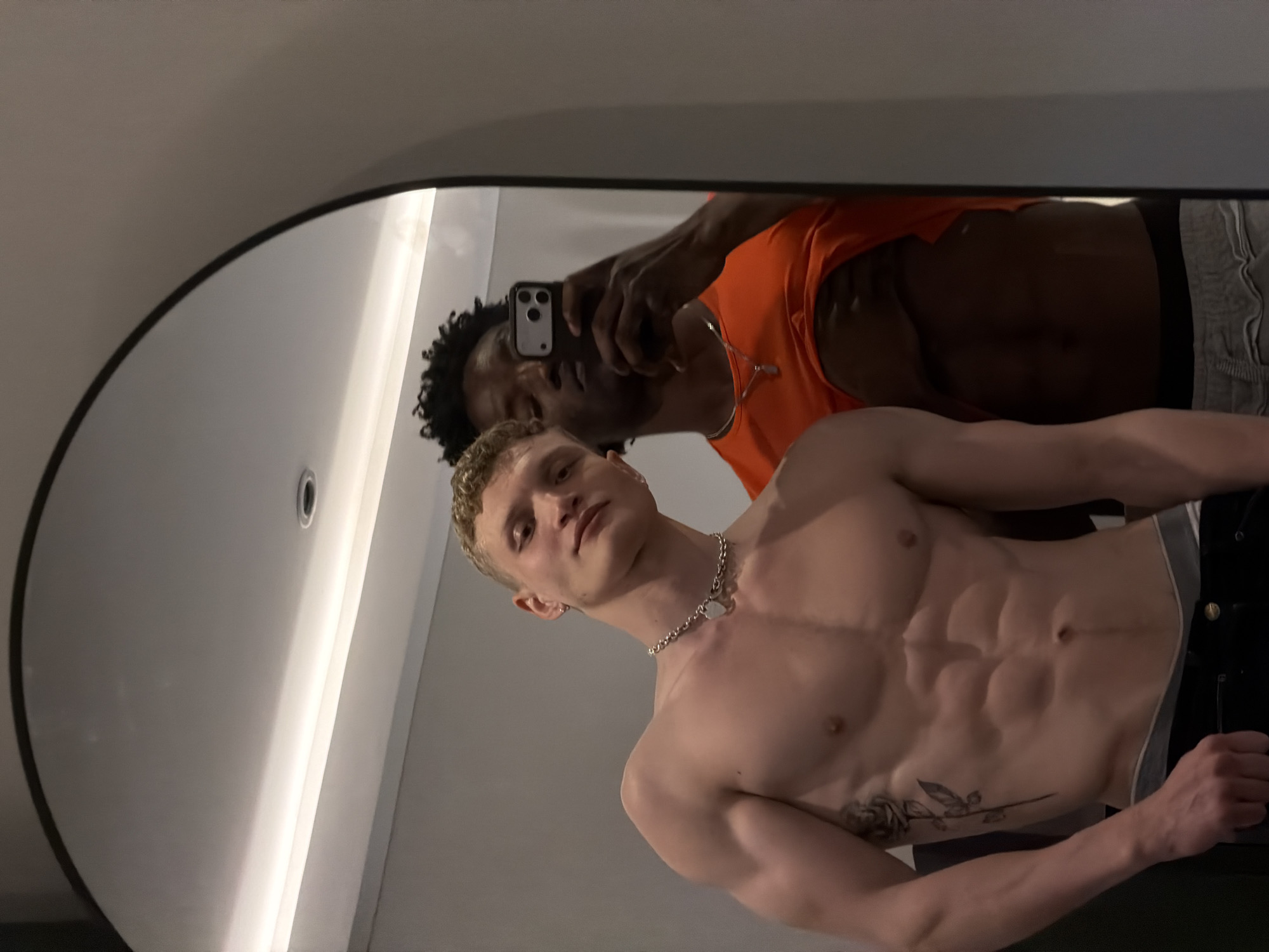 The image shows a selfie in a mirror. In the foreground, a shirtless, muscular white man with short blonde hair and a silver chain poses. A rose tattoo is visible on his torso. Behind him, an African-American man in an orange shirt holds a phone, presumably taking the picture. The background suggests an interior space with a light fixture on the ceiling. The location within London, United Kingdom is not discernible from the provided image.