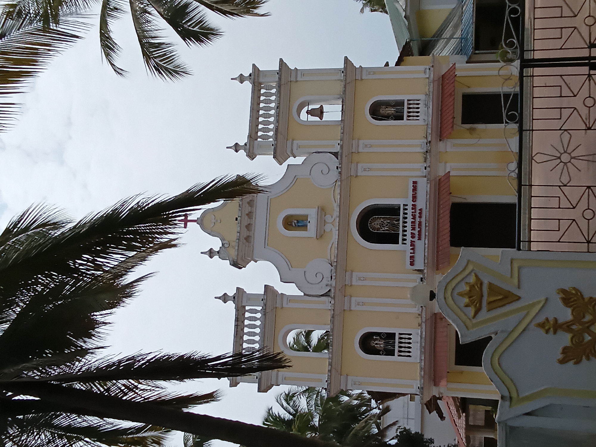 The image shows the Our Lady of Miracles Church in Ashvem, India, viewed from a low angle through palm trees. The church is a yellow and white building with a multi-tiered tower, with a bell visible in the top tier. Each tier has arched windows with decorative detailing. A sign reading "Our Lady of Miracles Church, Ashvem-Goa" is visible. To the right of the church, there is a fence or gate with a decorative pattern. The sky is overcast.