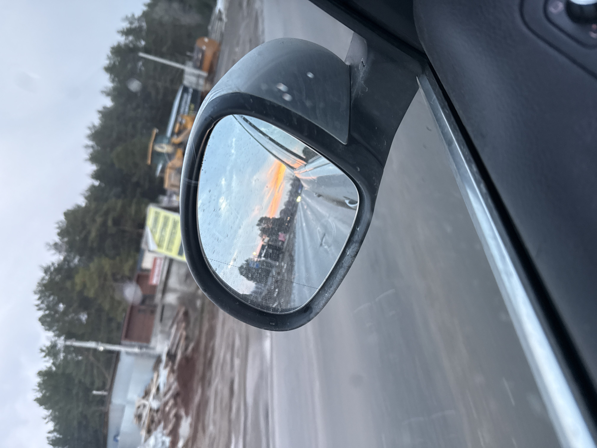The image shows a car's side mirror reflecting a road at sunset or sunrise. In the background, outside of the reflection, there are trees, buildings, construction equipment, and what appear to be piles of lumber or other materials. Raindrops are visible on the mirror. The image does not provide enough information to confirm the specific location as Chaikovsky, Russia, or any other location.