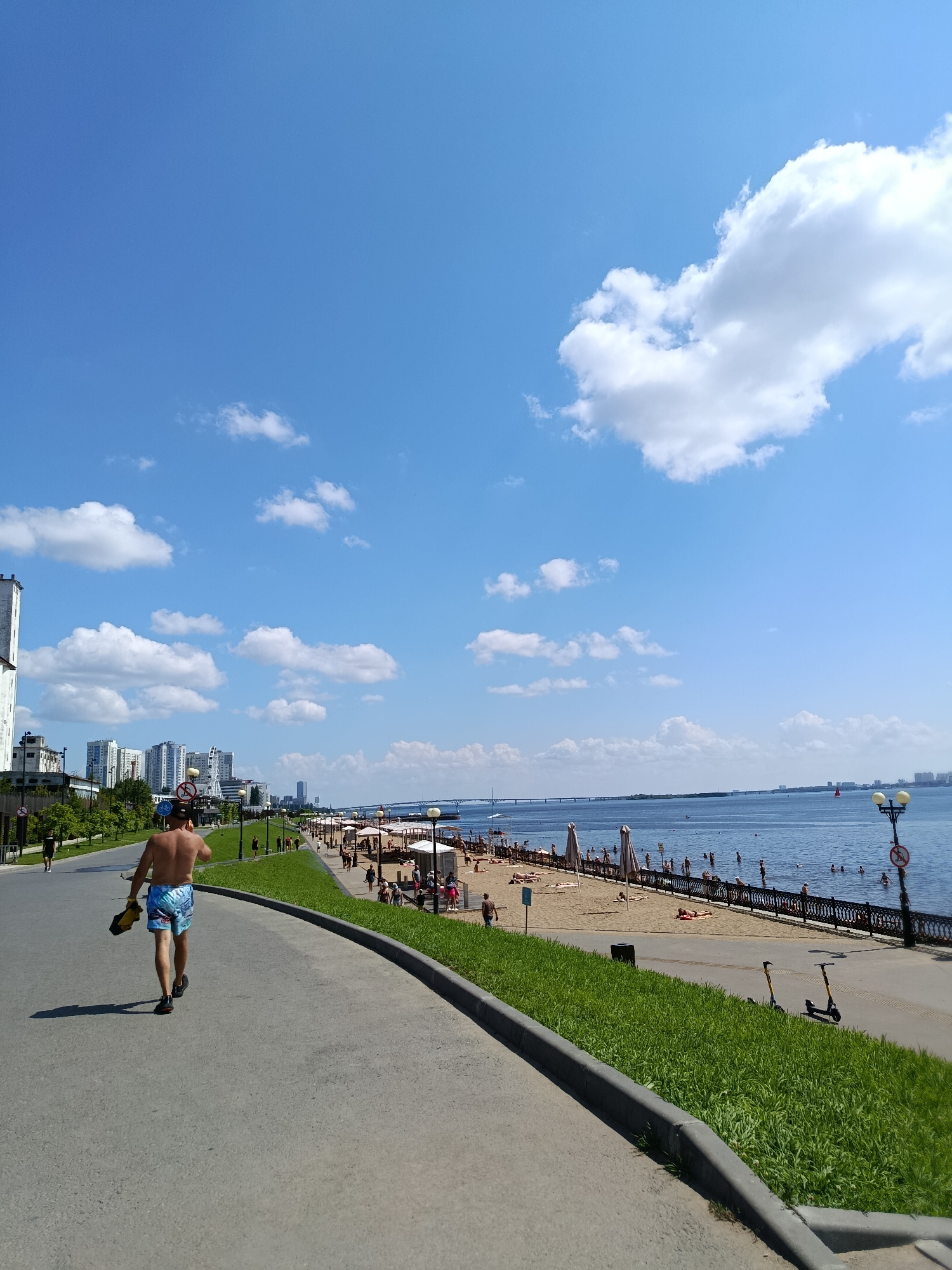 This image depicts a sunny day at a waterfront park and beach in Saratov, Russia. The scene is a vibrant summer day with a clear blue sky dotted with fluffy white clouds. The sun is high in the sky, indicating it is likely midday or early afternoon.
In the foreground, a person is walking away from the camera on a paved walkway. They are wearing blue patterned swim shorts and carrying a yellow object, possibly a bag or a mat. The walkway curves along the edge of a grassy embankment. Beyond the grass, a bustling beach area is visible, filled with people sunbathing on the sand, swimming in the water, and relaxing under beach umbrellas. The water of the wide river is calm and reflects the bright sky.
Along the edge of the beach, a decorative black fence separates it from the promenade, which is lined with lampposts and more people strolling. In the background, modern high-rise buildings and other structures suggest an urban environment adjacent to the recreational area. A long bridge spans the river in the distance.
The overall mood is one of leisure and enjoyment, characteristic of a warm summer day at a popular outdoor spot. The presence of swimsuits, beach umbrellas, and people in the water clearly indicates a beach setting where swimming and sunbathing are taking place. The image captures a typical summer scene in a city with a prominent riverfront. No visible text is present in the image.