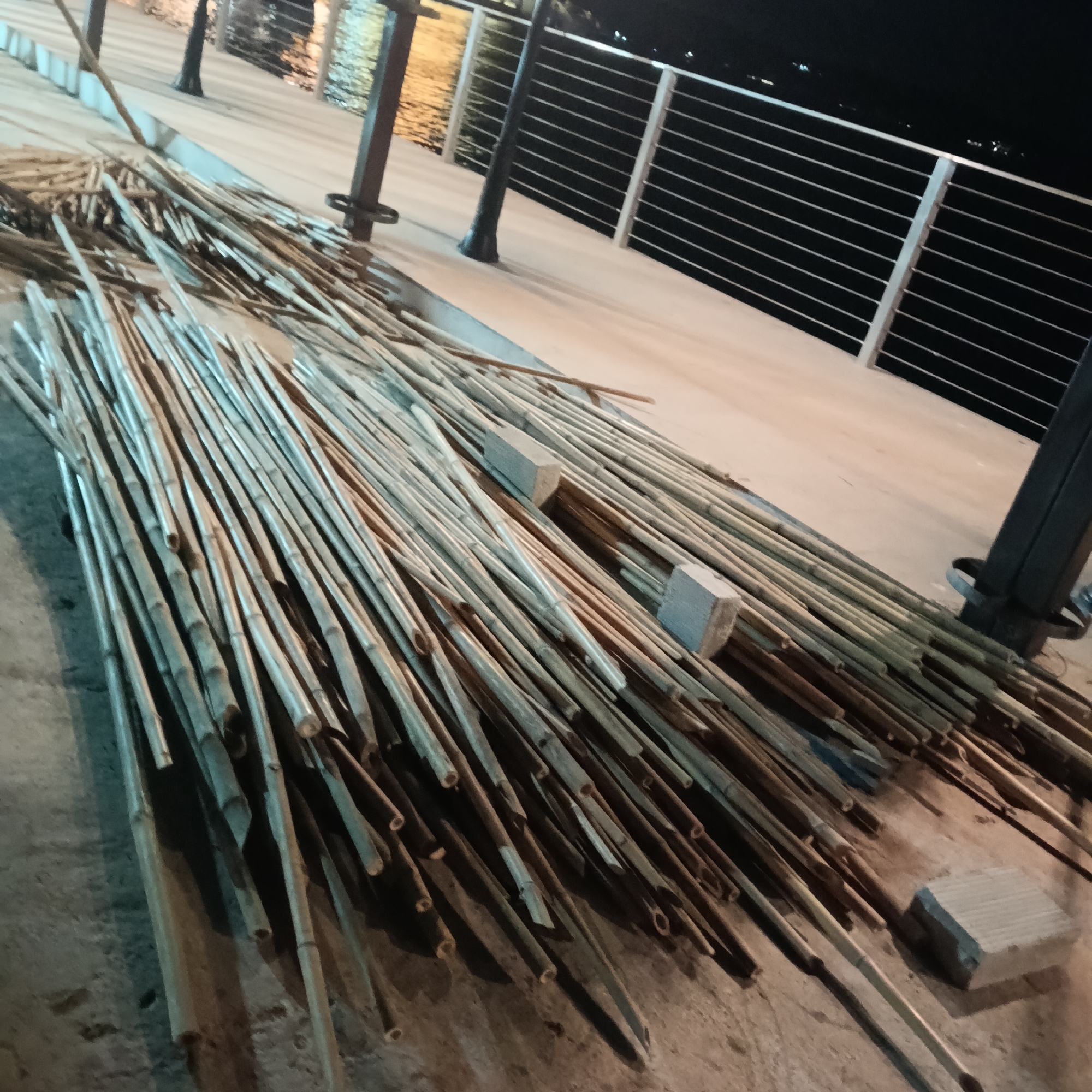 The image shows a collection of bamboo poles lying on a light-colored surface, possibly a pier or walkway. The poles appear to be of varying lengths and thicknesses. Some concrete blocks are interspersed among the poles, potentially used as supports or weights. In the background, there is a body of water reflecting light, and a railing runs along the edge of the pier. Lamp posts are visible along the railing. The scene is dimly lit, suggesting it is nighttime. Based on the presence of the Mandovi River, the location is likely near Panaji, India. No people are visible in the image.