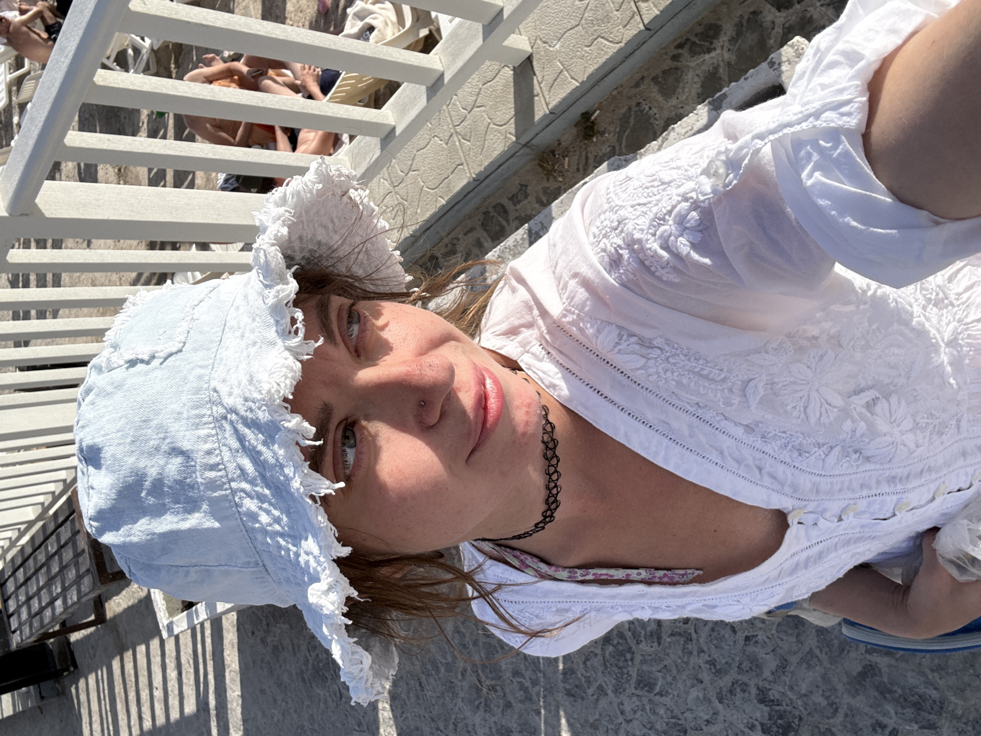 A woman is taking a selfie while sitting on a bench outdoors. She is wearing a light blue bucket hat and a white embroidered tunic. A black choker necklace is visible around her neck. The background shows a beach scene with lounge chairs and people in the distance. The ground is paved with light-colored stone. The lighting suggests it is daytime.