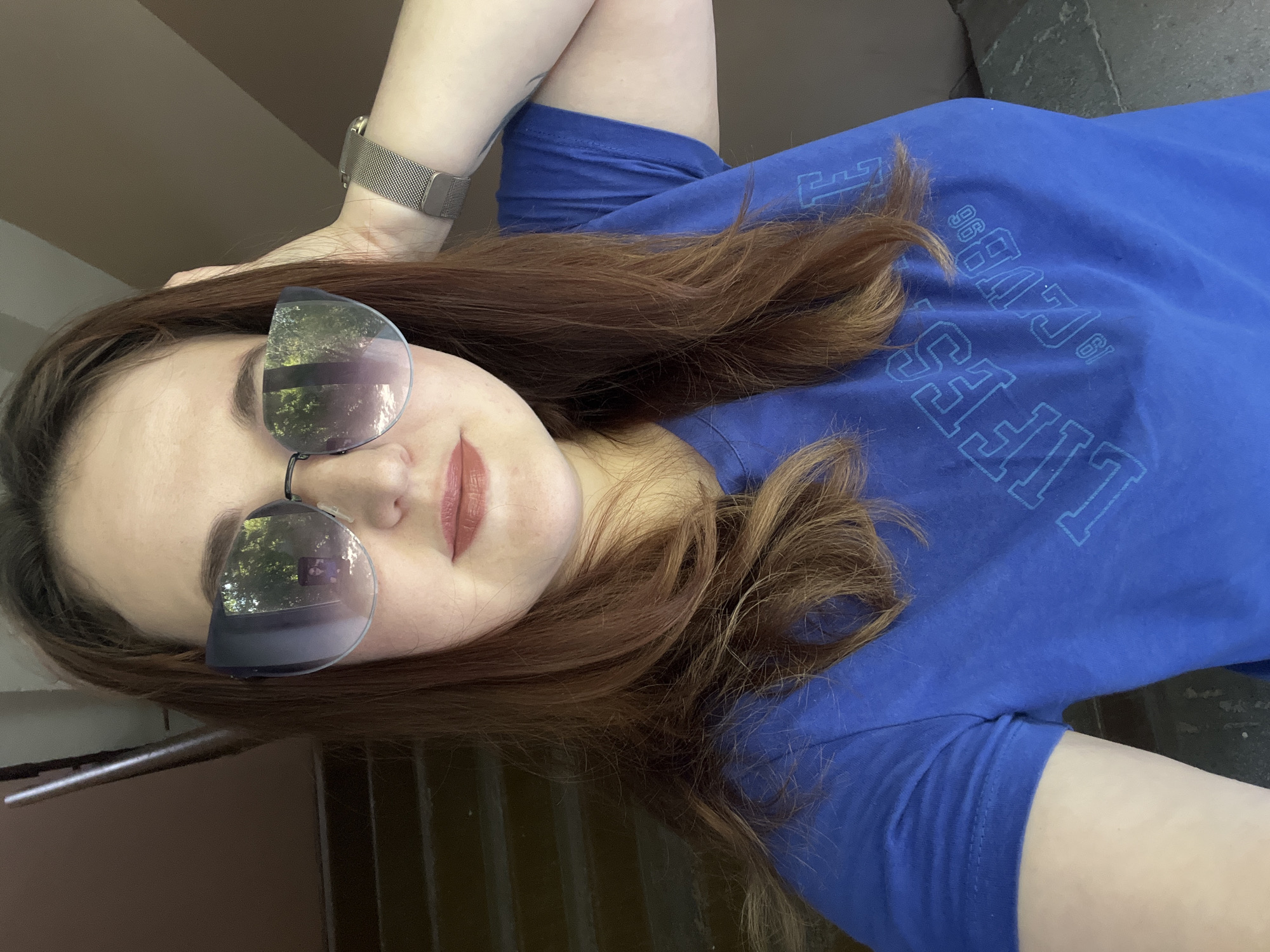 A young woman with auburn hair and light skin is taking a selfie. She is wearing a blue t-shirt with a graphic design and a silver watch on her left wrist. She has on large, stylish sunglasses that reflect a green, leafy outdoor scene. Her right hand is raised and touching the back of her head. The background appears to be a staircase with brown walls and railings. The woman's makeup includes a matte lipstick in a brownish-pink shade. The image has a slightly grainy quality.
