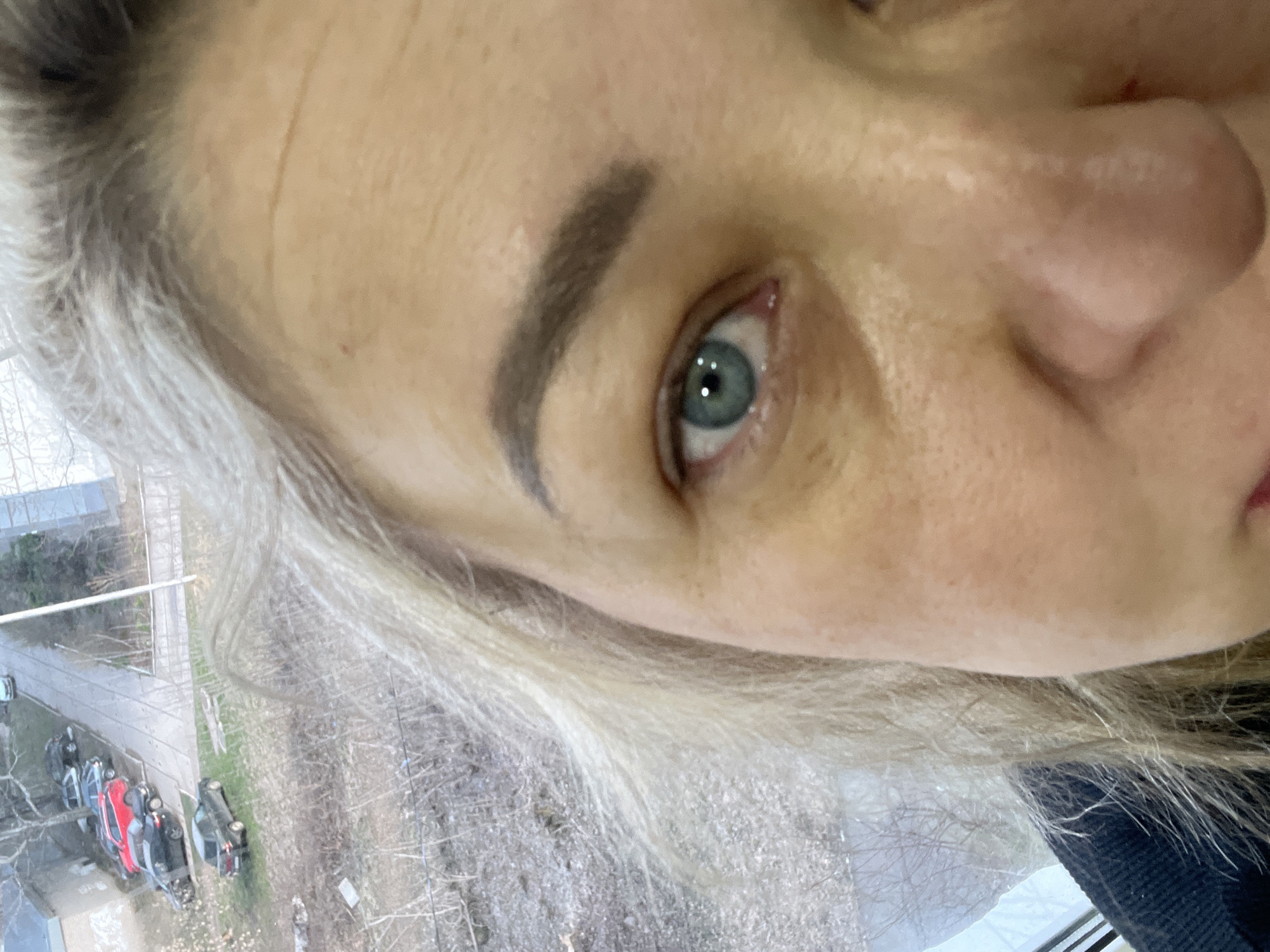 A close-up selfie of a woman with gray hair, fair skin, and blue eyes. A portion of her face is visible including her eyebrow, eye, nose, and part of her mouth. The image is taken at a slight angle. Outside the window, visible in the background, is a street with cars, including a red car. The location appears to be Chaikovsky, Russia, based on the car models and general environment visible through the window.
