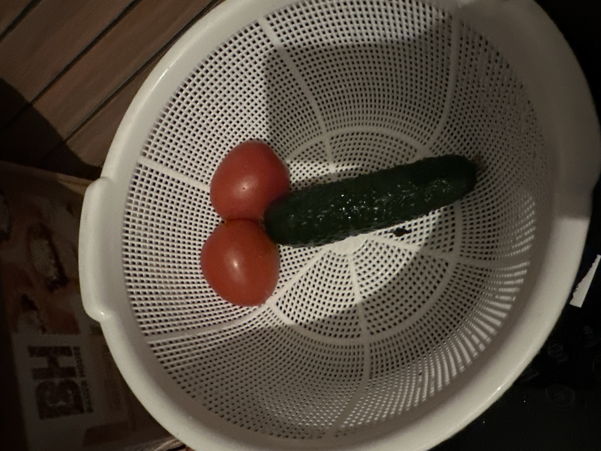 A white plastic colander containing two red tomatoes and one dark green cucumber is centered in the frame. The tomatoes are positioned side-by-side at the top, while the cucumber is placed vertically below them. The colander features a perforated design with numerous small square holes. In the upper left background, a brown package or object is partially visible, displaying the text "BH" and "BAKER HOUSE." The upper right background shows a dark brown surface with horizontal striations, resembling wood paneling. The lower background is dark and indistinct.