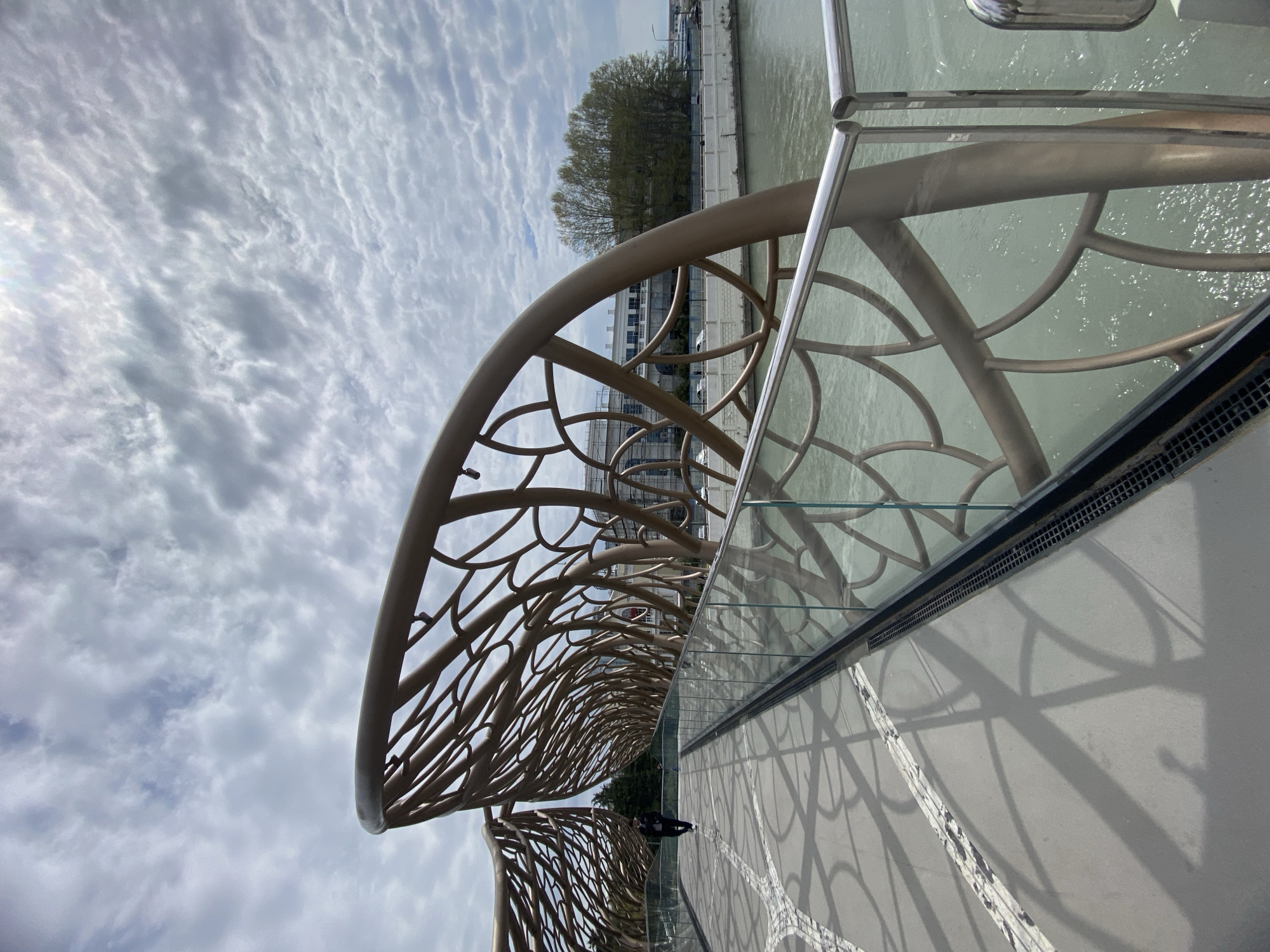 The image, taken in Sochi, Russia, captures a modern pedestrian bridge or walkway characterized by its unique architectural design. The most prominent feature is an ornate, light brown or bronze-colored metal structure that forms an arched canopy or balustrade along the pathway. This structure exhibits an intricate, organic pattern of interconnected branches and curves, reminiscent of veins or leaves.
The walkway is paved with light-colored tiles, some with a darker geometric pattern, and is adorned with complex shadows cast by the overhead structure, creating an artistic effect on the ground. A clear glass railing, topped with a metallic handrail, runs along the edge of the walkway, separating it from a body of water – likely a river or canal – which is visible on the right with a gently rippled surface. A drainage or ventilation grating runs along the base of the railing.
In the distance, a solitary person can be seen walking away from the camera on the bridge. Beyond the water, along the far bank, there is a line of trees with light green foliage, suggesting a springtime setting, and several buildings with light-colored facades. The sky above is partially overcast with scattered, textured clouds, indicating daytime conditions. There is no visible text in the image. The scene conveys a sense of contemporary design harmoniously integrated into the urban landscape.