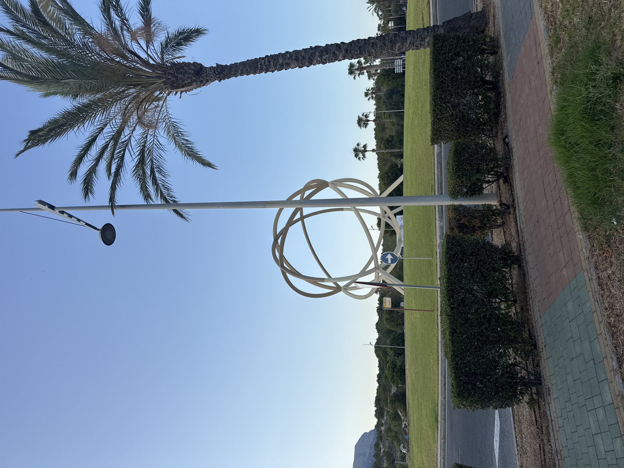 A large, modern sculpture composed of interlocking rings stands prominently in a grassy roundabout. A tall palm tree, partially obscuring a light pole, is situated to the right of the sculpture. In the background, lush green grass covers the roundabout, bordered by trees and a distant mountain range. To the left of the sculpture, a road curves around the roundabout, with a sidewalk and a row of neatly trimmed hedges in the foreground. The sky is a clear, pale blue, indicating a sunny day. This sculpture is located at the entrance to Benidorm, Spain, along the N-332 road.