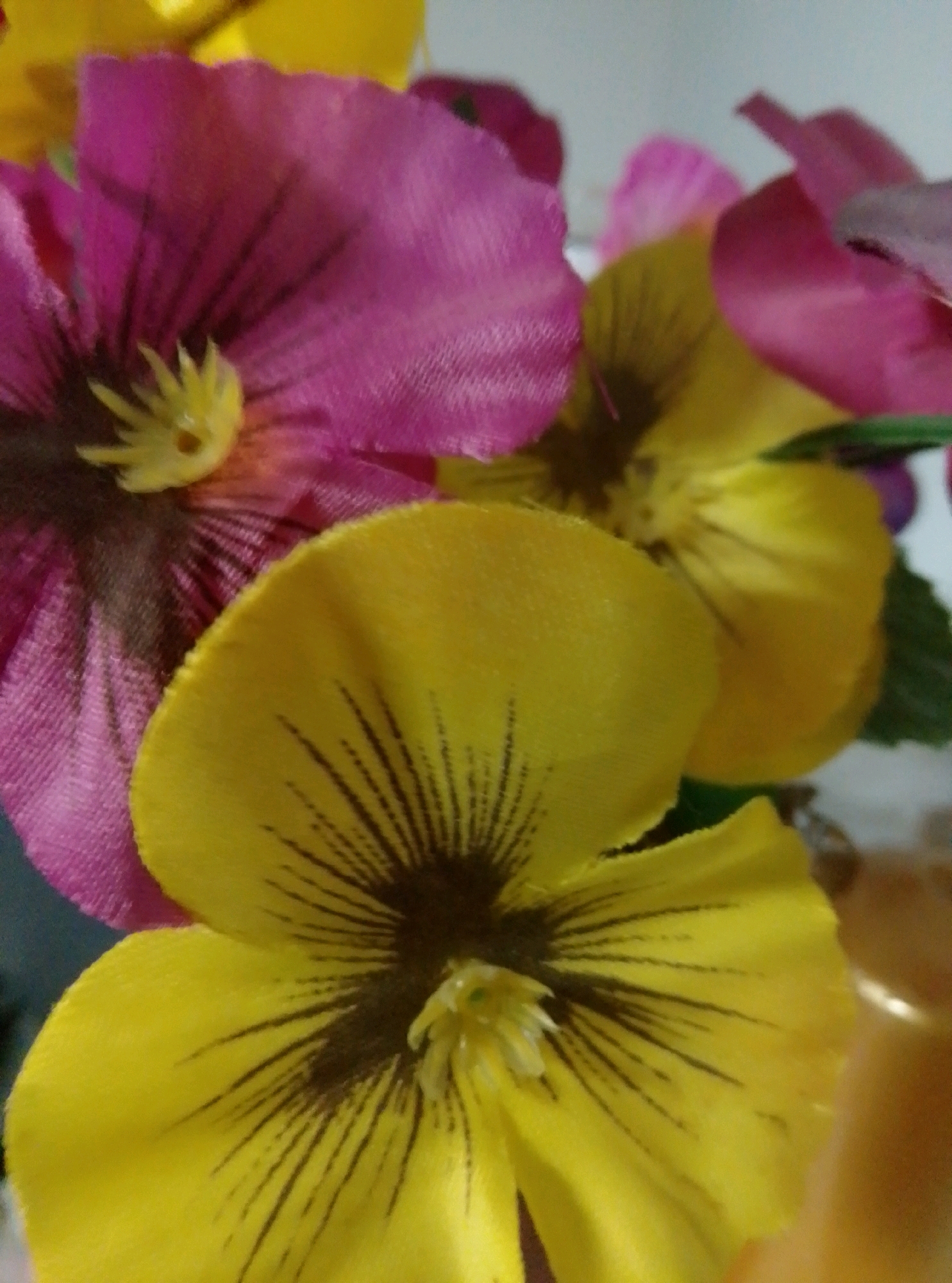 The image displays a close-up, low-angle view of several artificial pansy flowers. The flowers are vibrantly colored, with prominent yellow and pink petals, and dark centers with fine lines. The petals appear to be made of fabric. The background is blurred, suggesting a shallow depth of field. There are no people or actions depicted in the image. The setting does not provide any specific details about a location in the city of Wedel, Germany.