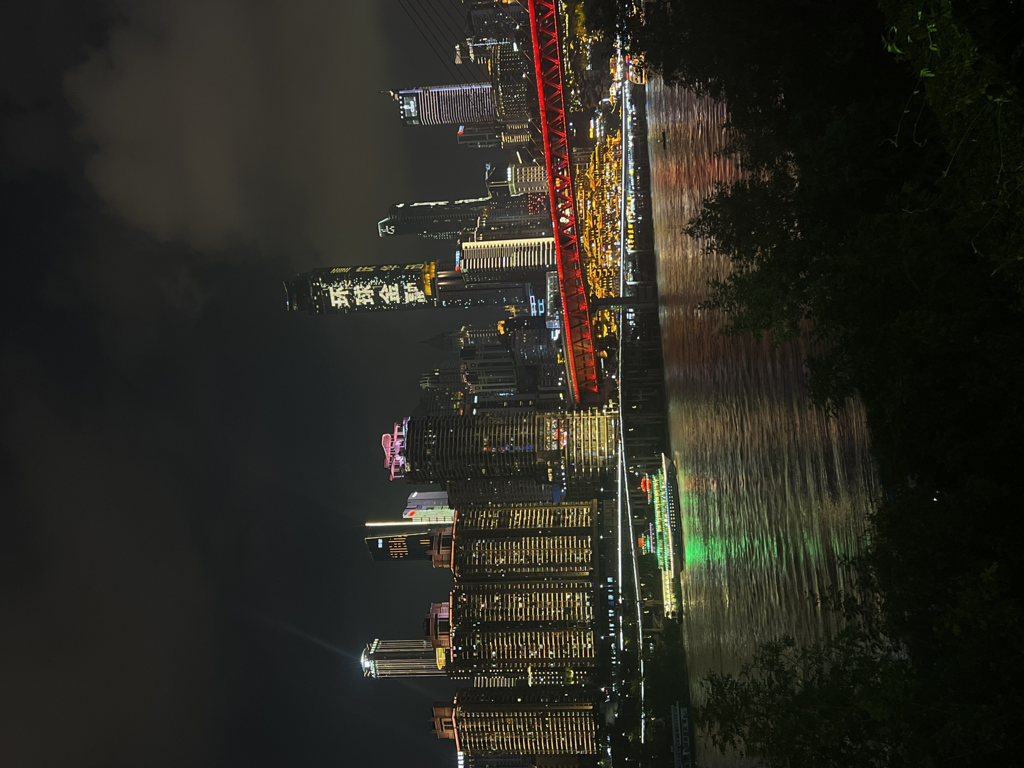 At night, the Jiangbeicheng Street area in China is illuminated by the city's skyline. A body of water, likely a river, flows in the foreground, reflecting the vibrant lights of the cityscape. On the water, a large, illuminated boat with green and red lights is visible, suggesting a leisurely cruise. Across the water, a modern city skyline dominates the background, featuring numerous skyscrapers of varying heights and architectural styles. Several buildings are lit up with warm yellow lights, creating a striking contrast against the dark night sky. One prominent skyscraper displays bright white characters, likely a prominent advertisement or landmark. A red illuminated bridge stretches across the water, connecting different parts of the city and adding a dramatic splash of color to the scene. The surrounding environment includes dark, silhouetted trees in the immediate foreground, framing the view of the city and the river. The sky is dark with some clouds, further emphasizing the illuminated city below.