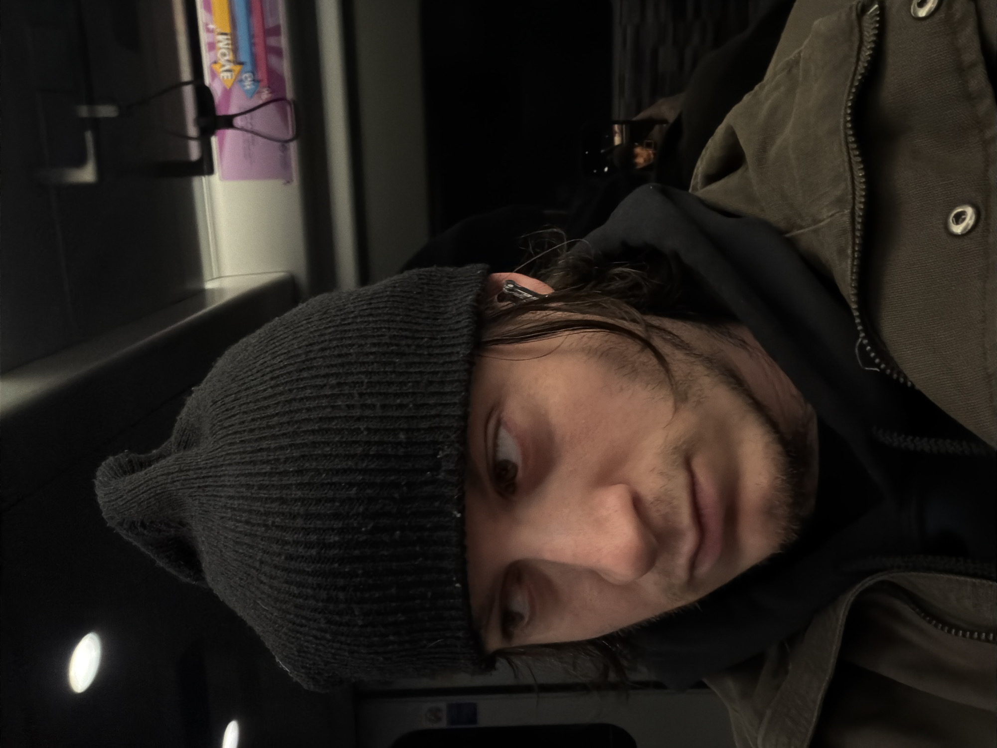 A close-up, rotated portrait shows a man wearing a dark gray knit beanie, a black hooded sweatshirt, and a brown, possibly waxed-canvas, jacket. The jacket is partially zipped. An earbud is visible in his ear. He appears to be seated inside a train carriage. A row of lights is visible in the background. A package of wet wipes or similar product is clipped to the carriage wall. The scene does not provide sufficient information to verify its location in London.