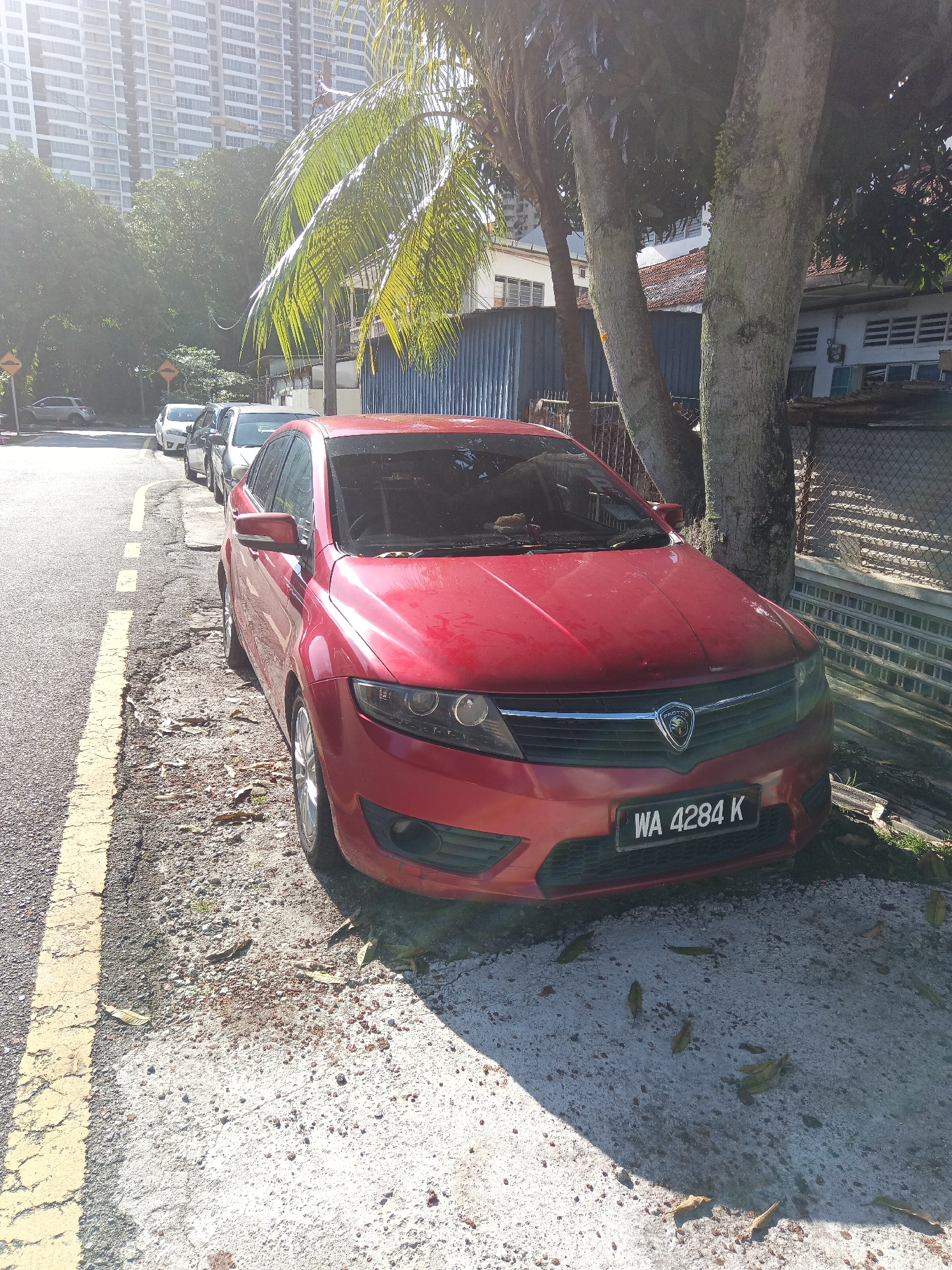 The image depicts a red Proton Prevé car parked on the side of a road in Kuala Lumpur, Malaysia. The car's license plate reads "WA 4284 K". The vehicle is positioned to the right of the frame, facing towards the viewer and slightly to the left. The road has a yellow double line marking on the left side, indicating parking is allowed. A tree with rough bark is situated directly behind the car, casting a shadow on the vehicle and the ground. In the background, a tall, modern apartment building is visible, suggesting an urban environment. Scattered dry leaves are present on the ground around the car. Other vehicles are parked further down the street, partially visible in the distance. The scene is illuminated by bright sunlight, creating strong contrasts and shadows.