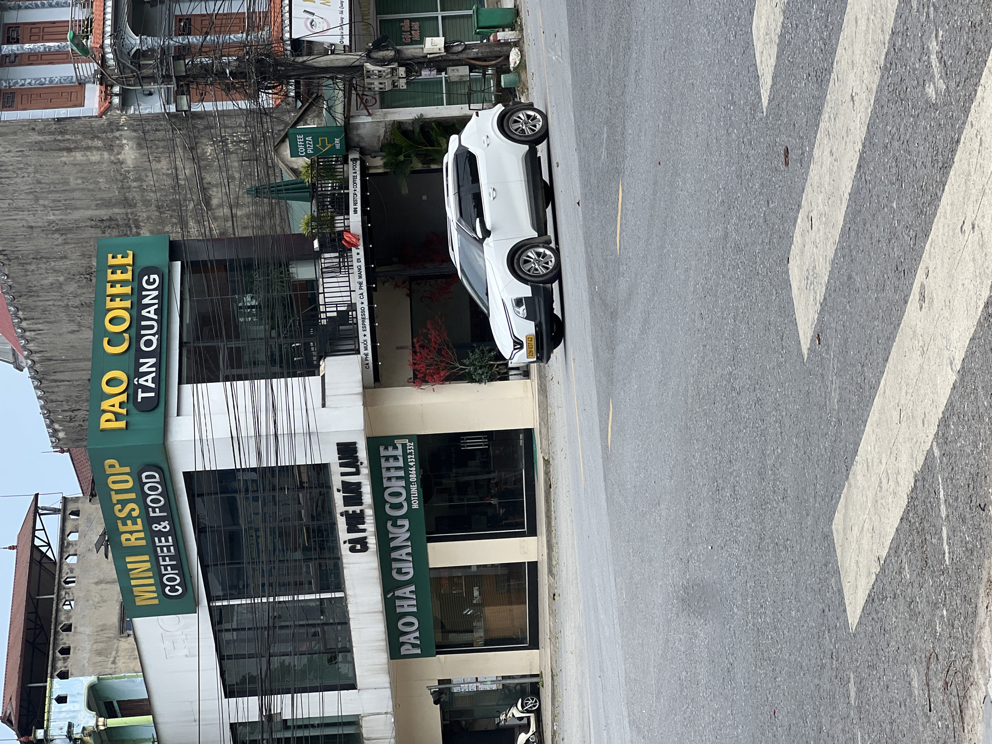 The image captures a street scene in Tan Quang Commune, Vietnam, featuring a white SUV parked on the right side of the road, partially obscuring a pedestrian crossing. The vehicle, identified by license plate "22H-037.42", is a modern crossover.
In the background, a multi-story building is prominently displayed, housing several businesses. The upper facade of this building features a large, green sign that reads "PAO COFFEE TAN QUANG", with smaller text above indicating "MINI RESTOP COFFEE & FOOD". Below this, another green sign for "PAO HA GIANG COFFEE" is visible, along with a hotline number. The lower levels of the building have large glass windows.
Adjacent to the coffee shops, there are other establishments, including one with a green sign pointing towards "COFFEE PIZZA HERE". A green awning and some potted plants are also visible. The street is paved and shows evidence of wear, with fallen leaves scattered on the asphalt and the pedestrian crossing. The sky is overcast. The scene is typical of a commercial area in a Vietnamese town, with a mix of modern businesses and older architecture, and the ubiquitous presence of electrical wires overhead.