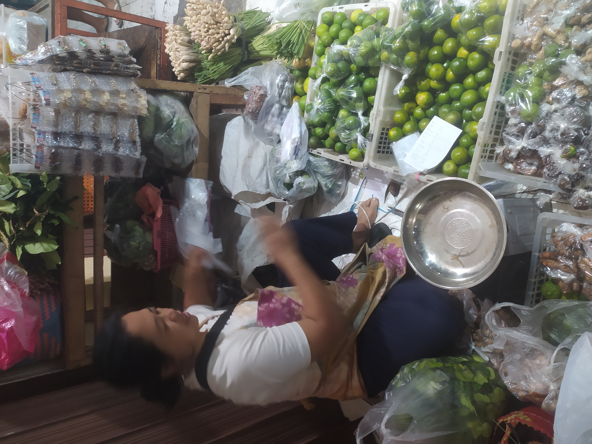 A medium shot captures a female vendor seated in front of a market stall filled with produce and packaged goods. The vendor, wearing a white t-shirt and a patterned apron, is actively engaged in handling what appears to be plastic bags of produce. Her facial expression is neutral, and her hands are in motion, suggesting she is in the process of bagging or preparing items for sale.
The stall itself is laden with a variety of goods. In the foreground, several plastic crates overflow with green citrus fruits, possibly limes, some of which are individually bagged. Adjacent to these crates are other items, including what seem to be bags of spices or roots, and smaller packaged goods. In the background, more produce is visible, including bunches of what looks like lemongrass or similar stalks. Shelving units behind the vendor hold more packaged items, some hanging from racks, and baskets containing other produce. The overall environment suggests a bustling local market. The lighting is dim, and the overall impression is one of a functional, working marketplace. The scene is consistent with a typical market setting found in Kupang, Indonesia, where such stalls are common for vendors selling fresh produce and local goods.