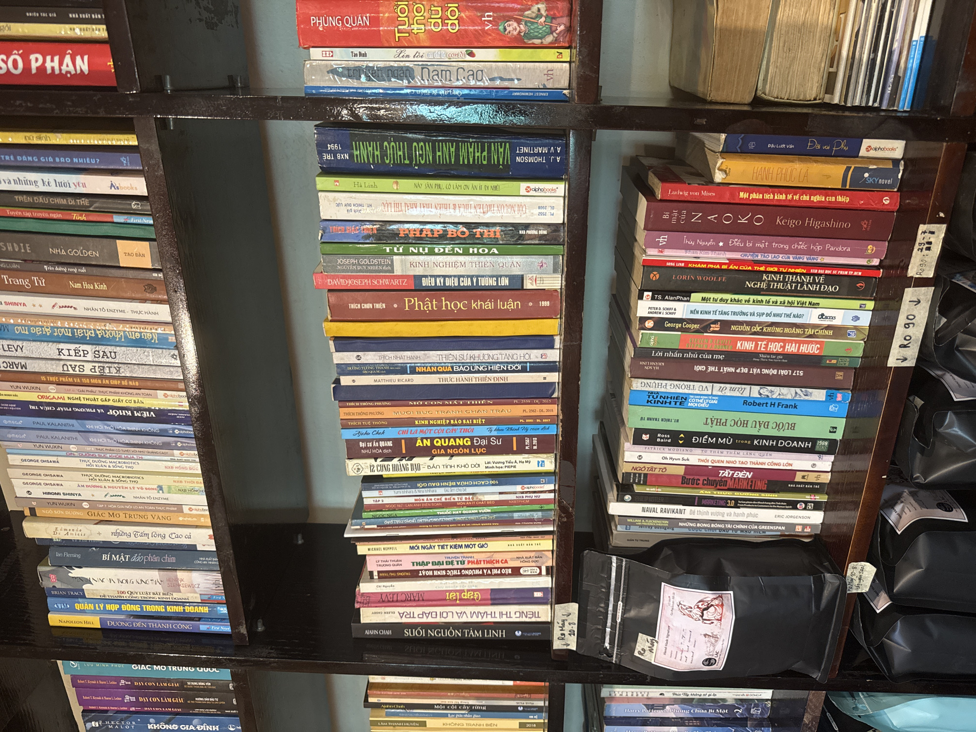 The image shows a close-up, eye-level view of several shelves of books in what appears to be a cafe or bookstore. The shelves are dark wood, and the books are arranged vertically, spine out. The books are a mix of hardcover and paperback, with a variety of colors and sizes. Some of the books have Vietnamese titles, such as "Sổ Phận," "Tựa Dõi," and "An Quang Đại Sự." There is also a black bag of coffee beans with a label that says "Ro May" and "Hand-Made Signature." The overall impression is of a cozy, well-stocked space with a literary theme. It is not possible to determine the exact location in Phú Thủy, Vietnam from the image alone.