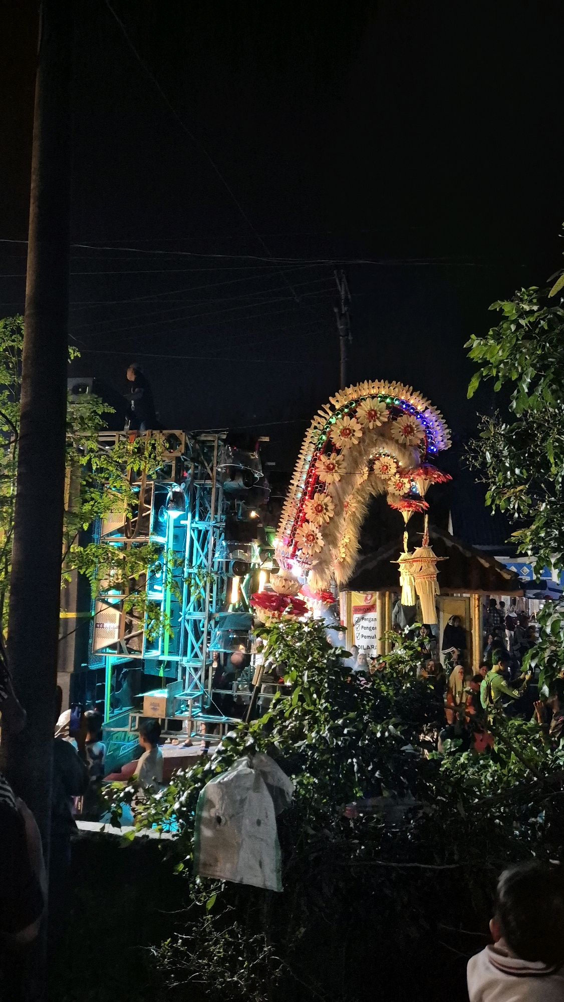 A night scene in Tanjungarum, Indonesia, features a vibrant, illuminated dragon float decorated with flowers and lights. The float is positioned in front of a large sound system stacked with speakers, illuminated by blue lights. A person stands on top of the sound system. In the background, a tent-like structure with signs and people milling about suggests a public event or gathering. Dense foliage partially obscures the lower portion of the scene, with a white plastic bag hanging from a branch. The sky is dark, indicating nighttime.