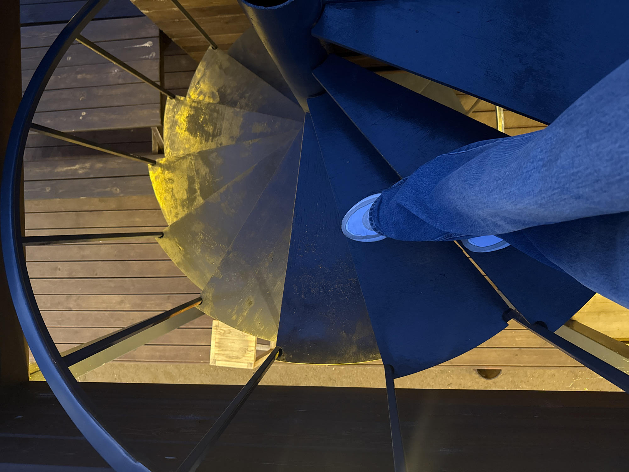 An individual's legs are visible, clad in blue jeans and white sneakers, as they descend a dark, spiral staircase. The staircase has metal treads, some of which appear worn and discolored, contrasting with the dark railing. The background consists of wooden planks, illuminated by warm, artificial light, suggesting an outdoor or semi-outdoor setting, possibly a deck or mezzanine. The image does not contain any discernible landmarks or features that would allow for identification of its location within Votkinsk, Russia.