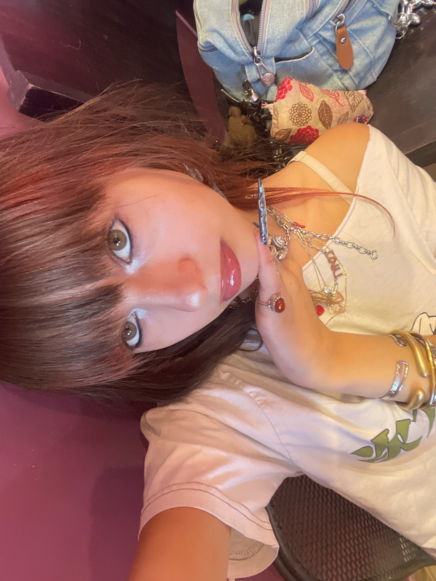 An individual with reddish-brown hair and bangs is lying down and looking at the camera. They are wearing a white t-shirt with a green graphic on it. The individual has makeup on, including eyeliner and lip gloss. They are adorned with several pieces of jewelry, including rings, necklaces, and bracelets. One of the rings on their left hand has an orange stone. The background is out of focus but appears to be a flat surface with some bags and other objects. The lighting suggests an indoor environment. The image does not contain any identifiable landmarks or features that would allow for its location to be determined within Raanana, Israel, or any other specific city.
