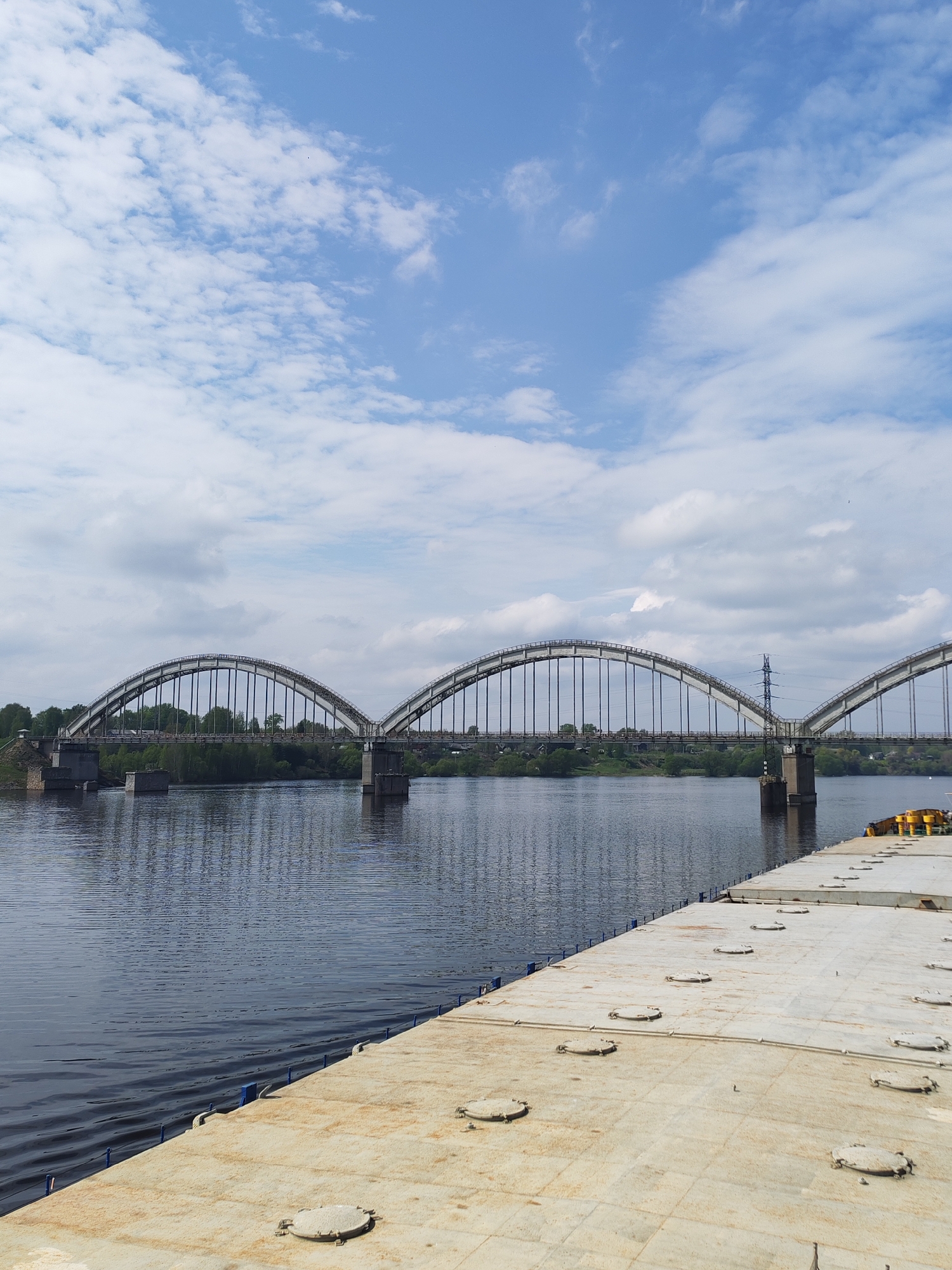 The image depicts an arched bridge spanning a body of water. The bridge has multiple arches supported by pillars in the water. The arches are connected by vertical supports. Green vegetation is visible on the banks of the water. Overhead power lines run near the bridge. The sky is blue with scattered white clouds. In the foreground, there is a concrete platform or dock with circular hatches. The overall location appears to be a riverfront area, possibly in the city of Zarya, Russia.