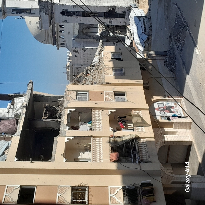 The image displays a street scene in Khan Yunis, Palestinian Territory, featuring severely damaged urban structures under a clear blue sky. In the foreground, multiple utility cables diagonally cross the frame. On the left, a multi-story, light-beige residential building exhibits significant structural damage on its upper two floors, revealing exposed rebar and missing concrete sections. Lower balconies, adorned with white balustrades, have laundry and tarpaulins hanging. A ground-floor shop entrance displays a sign for "YASEEN GAMES" with geometric graphics; a child wearing a light-colored shirt and a blue backpack stands near this entrance. To the right of this building, a large pile of rubble indicates a collapsed structure, with a white tent-like shelter positioned adjacent. In the background, a light-colored mosque, identified by its dome and minaret, also shows substantial damage, including pockmarks and partial structural failure on its walls. Debris piles are visible on the street and surrounding the damaged buildings. Strong sunlight casts distinct shadows across the scene.