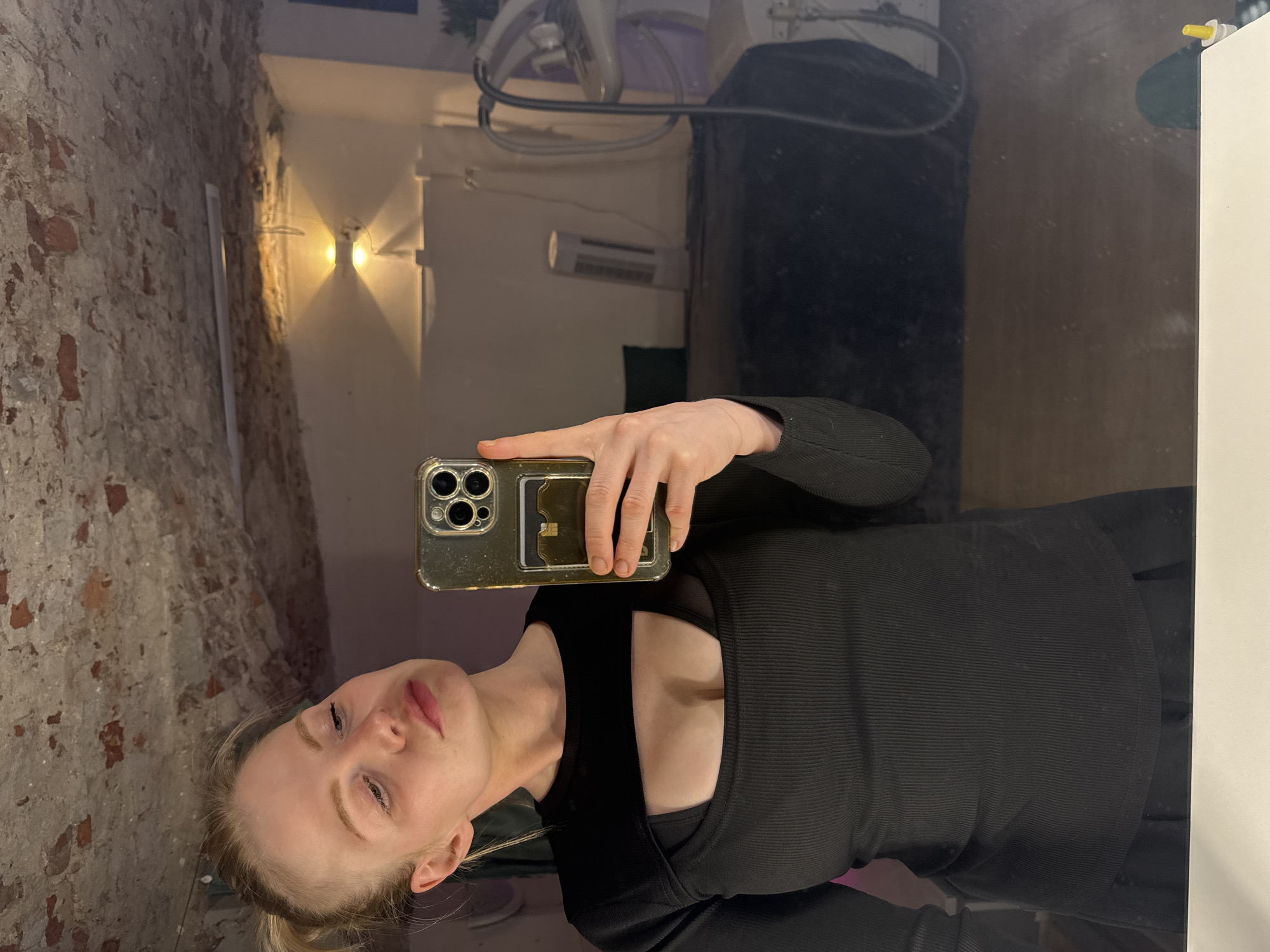 A young woman takes a mirror selfie in what appears to be a bathroom or dressing room. She is fair-skinned, with blonde hair pulled back, and is wearing a black, fitted top with a cut-out neckline. She is holding a smartphone, likely to capture the image. The background is a reflection in the mirror. The reflection shows a wall with exposed brickwork, suggesting a building with older architecture. There is a white wall and a drying machine. Given the information, the location could be in Saint Petersburg, Russia, known for its historical buildings and refurbished interiors with exposed brick.