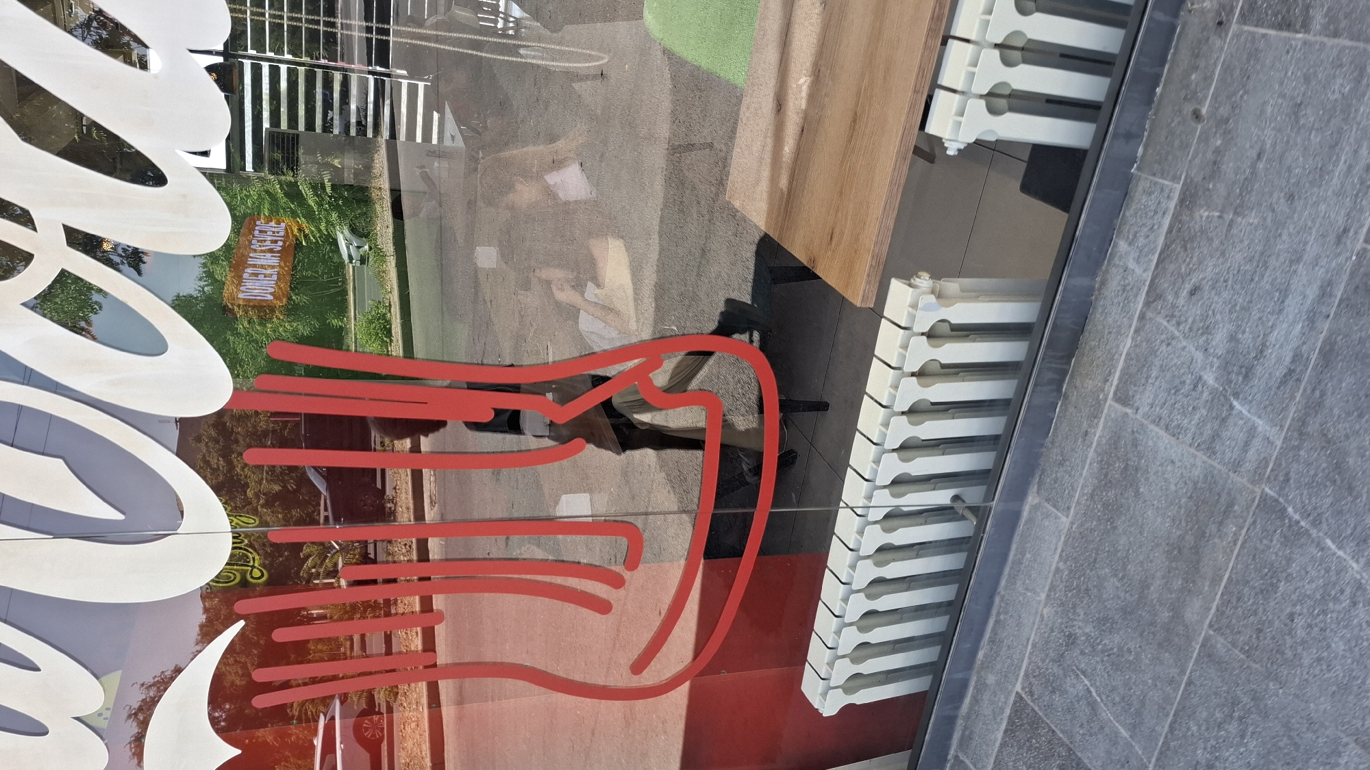 The image shows a reflection in a glass window of a street scene. Prominently featured are large white cursive letters that appear to be part of a shop's signage. In the reflection, there is a person standing, partially obscured by red vertical graphics on the window. Behind the person, a street is visible with trees and cars. A sign with the text "DONER NA SEVERE" is also visible in the reflection. Inside the building, hinted at by the reflection, are tables and chairs. In the foreground of the image, outside the window, are two white radiators. The ground is paved with grey tiles. The overall impression is that of a commercial establishment, likely a restaurant or cafe, located on a street. The presence of the "DONER NA SEVERE" sign suggests a food service business. The image is captured during daylight. This scene is consistent with an urban environment in Shymkent, Kazakhstan, where such commercial establishments and streetscapes are common.