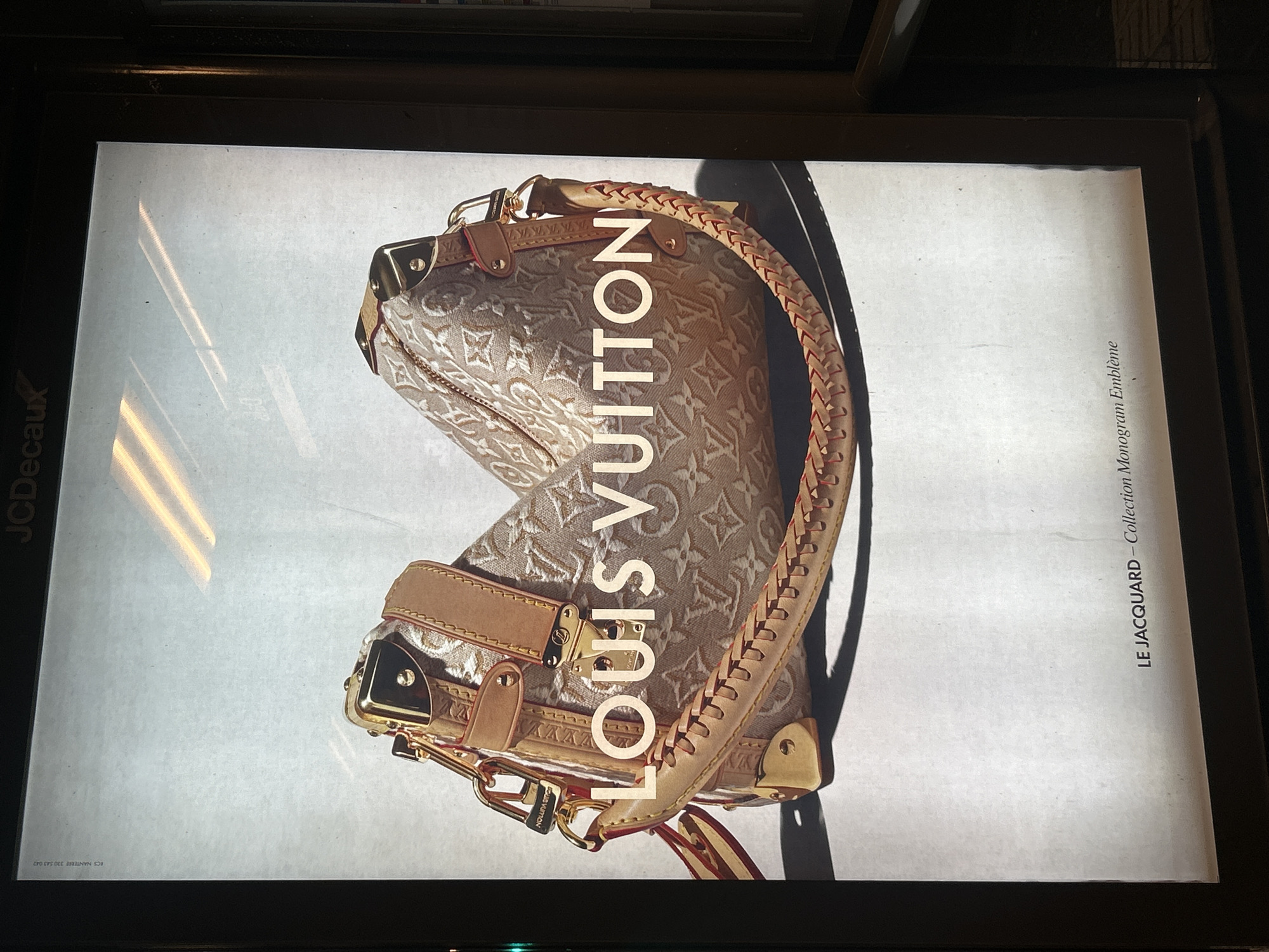 The image shows an advertisement for Louis Vuitton featuring a beige handbag with the brand's signature monogram pattern. The text "LOUIS VUITTON" is prominently displayed across the center of the bag. Below the bag, smaller text reads "LE JACQUARD - Collection Monogram Emblème." The advertisement appears to be displayed in a backlit public space, likely a bus shelter or similar outdoor advertising structure, as indicated by the "JCDecaux" logo at the top. The background is a plain, lightly textured white surface with two horizontal light fixtures visible at the top. The overall impression is of a clean and upscale product advertisement. The location is specified as a JCDecaux advertisement panel in Strasbourg, France.