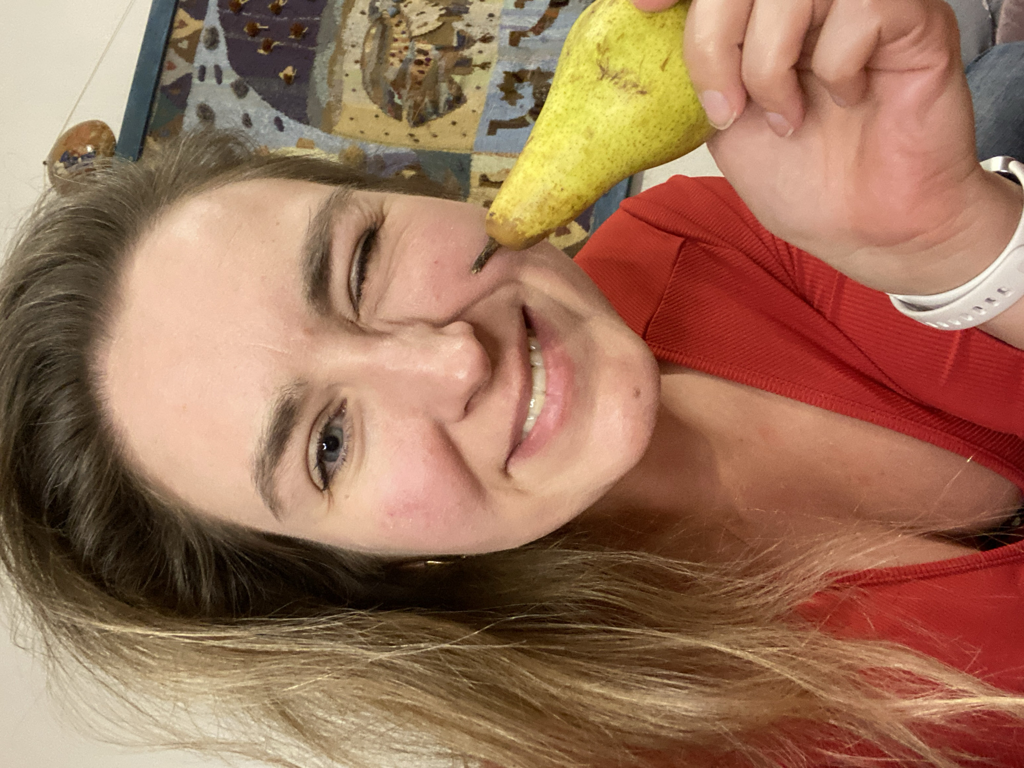 A woman is holding a pear to her face, specifically touching the tip of the pear to her cheek. She is smiling and winking. She is wearing a red shirt and a white watch. In the background, there is a patterned tapestry. The location is unknown and cannot be assumed to be in Yekaterinburg, Russia, based on the image alone.
