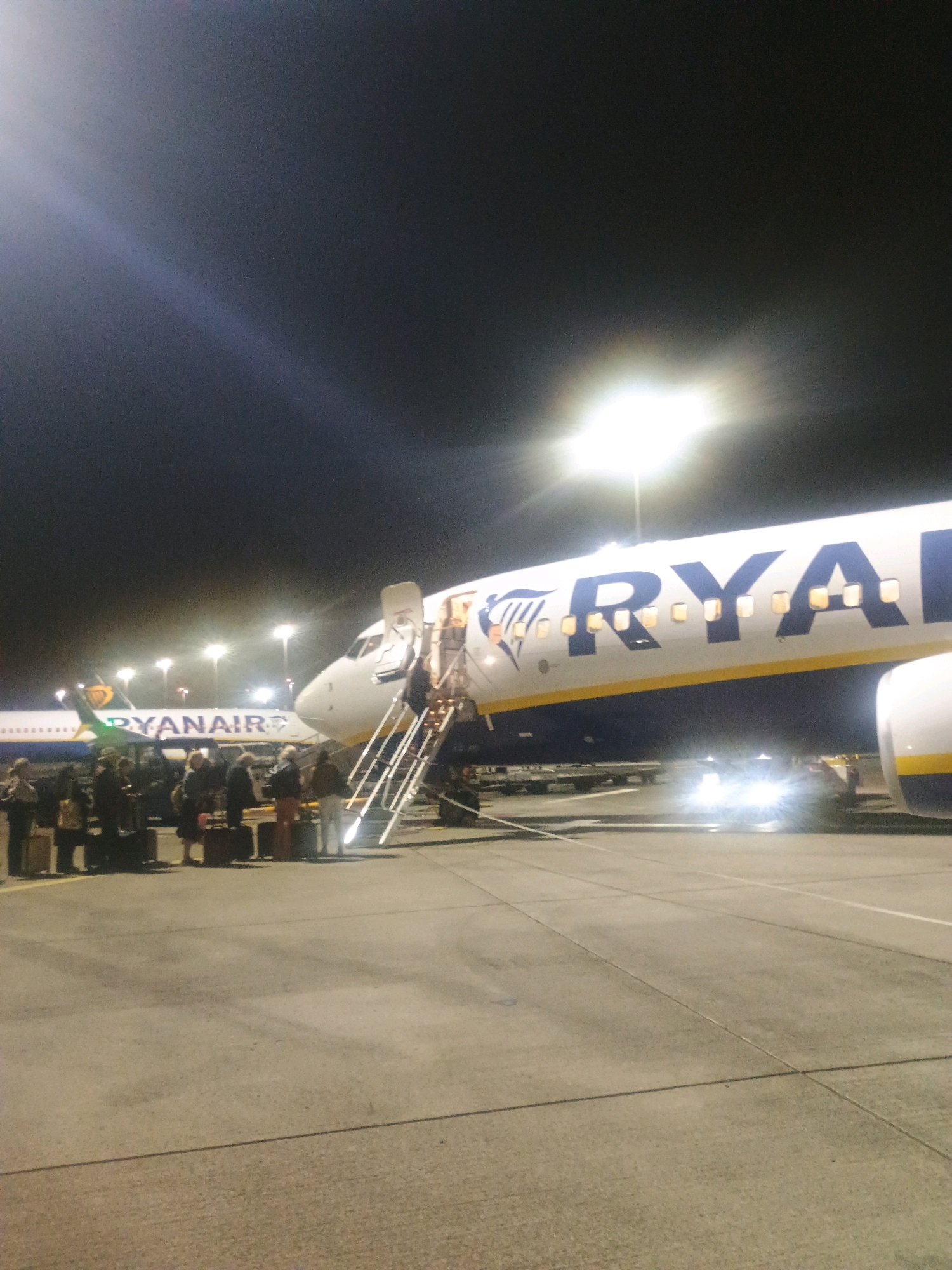 At night on an airport tarmac, two Ryanair airplanes are visible. The plane on the right is closer to the camera, its front section and partial fuselage illuminated by bright lights. A set of stairs leads up to an open door on the right side of the aircraft. Several passengers are gathered on the tarmac near the base of the stairs, some with luggage. The second Ryanair plane is further in the background on the left, its tail and a portion of its fuselage clearly visible. The sky above is dark, with several airport lights illuminating the scene. The ground is paved asphalt.