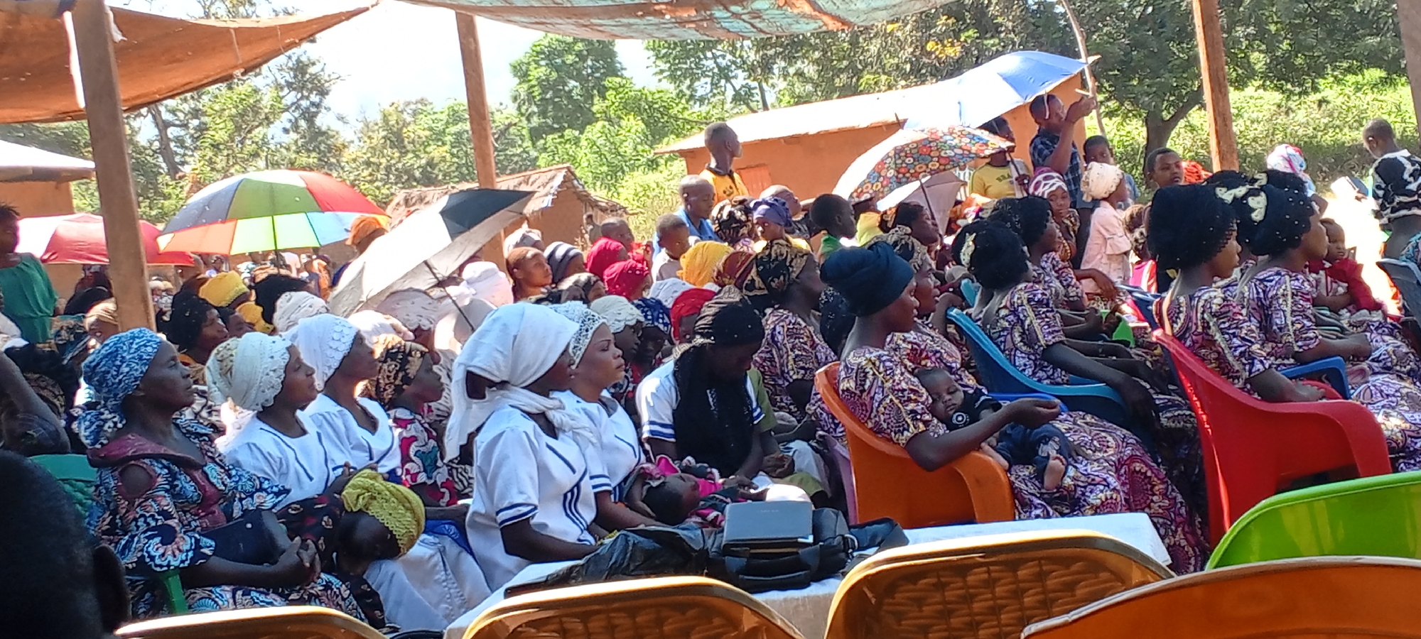 The image depicts a gathering of people, predominantly women, seated outdoors under a makeshift canopy. The location appears to be in a rural or semi-rural area, possibly near Ngalula, Democratic Republic of the Congo, given the description.
Many of the women are wearing similar brightly patterned dresses and head coverings, while others are in white uniforms with blue trim. Several individuals are holding babies or small children. Some umbrellas are open, providing additional shade.
The seating arrangement consists of rows of individual plastic chairs in various colors. In the foreground, a table is visible with equipment on top, including a bag and a possible electronic device. In the background, simple buildings and trees are visible. The scene suggests a community event or religious gathering.