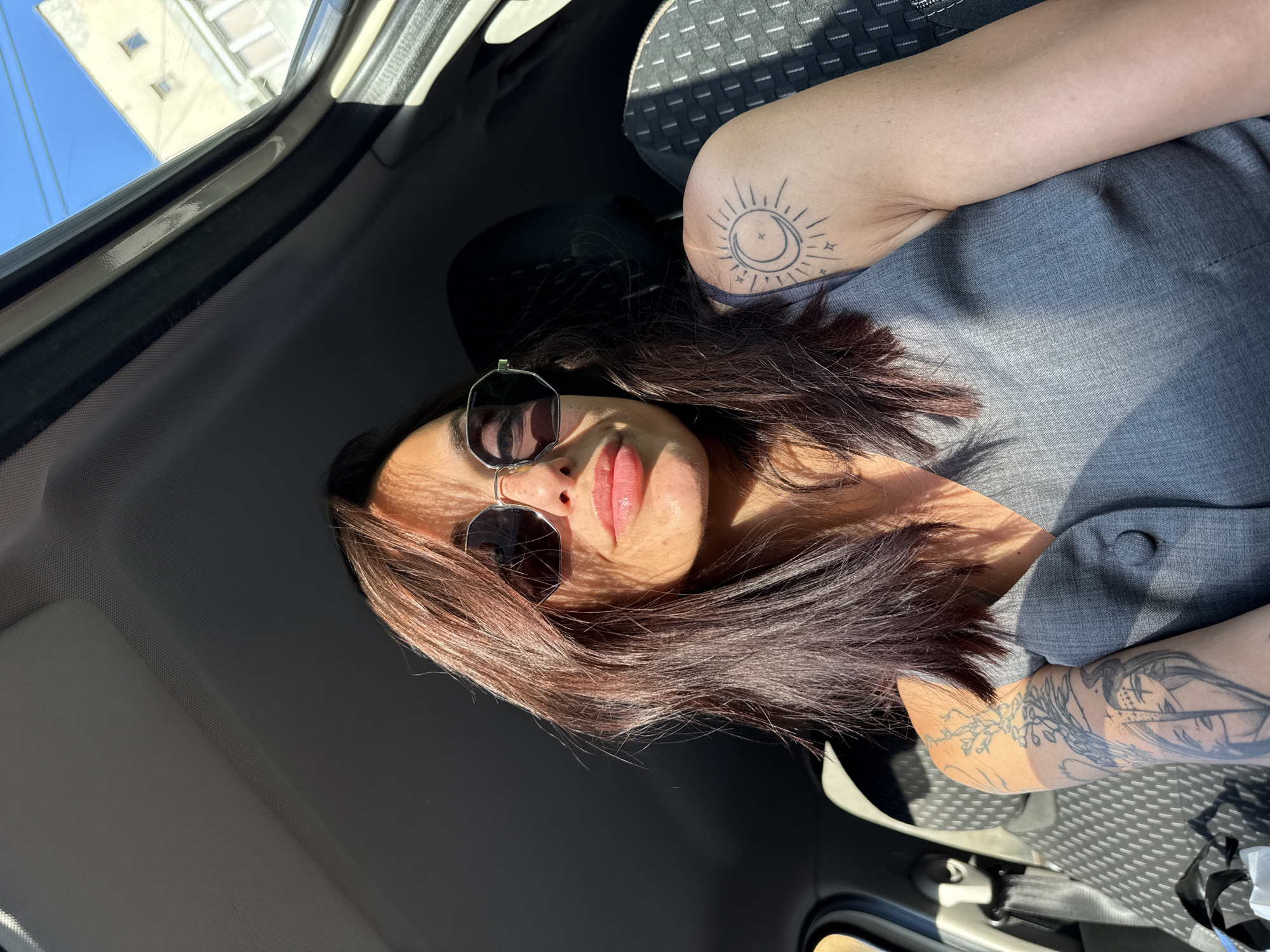 A medium shot shows a woman with auburn hair and sunglasses sitting in a car. The woman is in the passenger seat. She is wearing a grey shirt. There is a tattoo of the sun on her right arm. The photo was taken inside a car, with the building visible in the background. Based on the architecture and the license plate, the location is Pleven, Bulgaria.