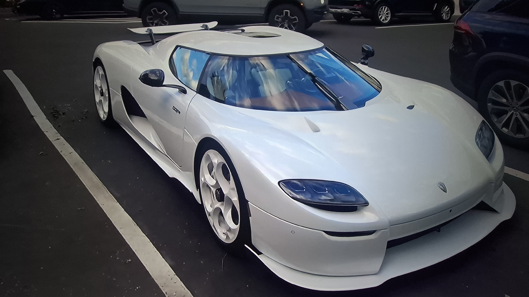 A white Koenigsegg supercar is the central subject of the image, parked in what appears to be an outdoor parking lot or street. The car is angled slightly, showcasing its front and driver's side. It features a sleek, aerodynamic design with distinctive multi-spoke white wheels, a prominent rear wing, and a single windshield wiper. The headlights are dark-tinted and integrated into the smooth front fascia. Reflections of a blue sky and white clouds are visible on the car's glossy surfaces, particularly the windshield and roof, indicating it is daytime. The interior, visible through the windshield, appears to have light-colored seats. A small badge with indistinct text, possibly "CCX" or similar model designation, is present on the side panel below the side mirror.
The setting consists of dark asphalt pavement with a single white parking line running alongside the car on the left. Scattered small debris, possibly leaves or dirt, is visible on the asphalt near the line. In the background, several other vehicles, predominantly dark-colored SUVs or sedans, are parked or driving. The lighting suggests a bright, possibly sunny or partly cloudy, day. No people are visible in the scene. The overall impression is of a high-performance vehicle stationary in an urban or suburban parking environment.