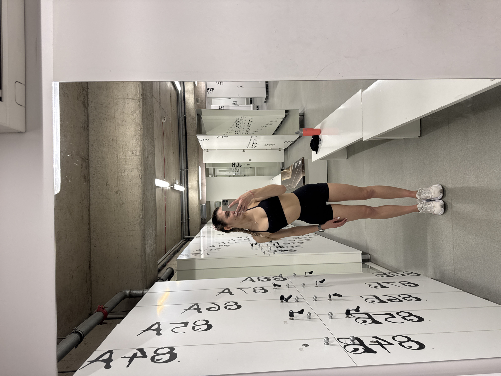 A young woman in athletic wear is taking a selfie in a locker room. She is wearing a black sports bra and black shorts, with white sneakers. She is holding her phone up to take the picture, facing the mirror. The locker room has rows of white lockers with black numbering. The lockers are in a well-lit space with a concrete wall visible in the reflection. There is a white bench beside the lockers. The OCR text includes "UTI," "Apr," "Err," "Srr," "ELLE," "orr," "AHVAD," "AT8," "ADS," and "AF8," which seem to be locker numbers. No specific details point to the city of Sofia, Bulgaria, so that cannot be confirmed.