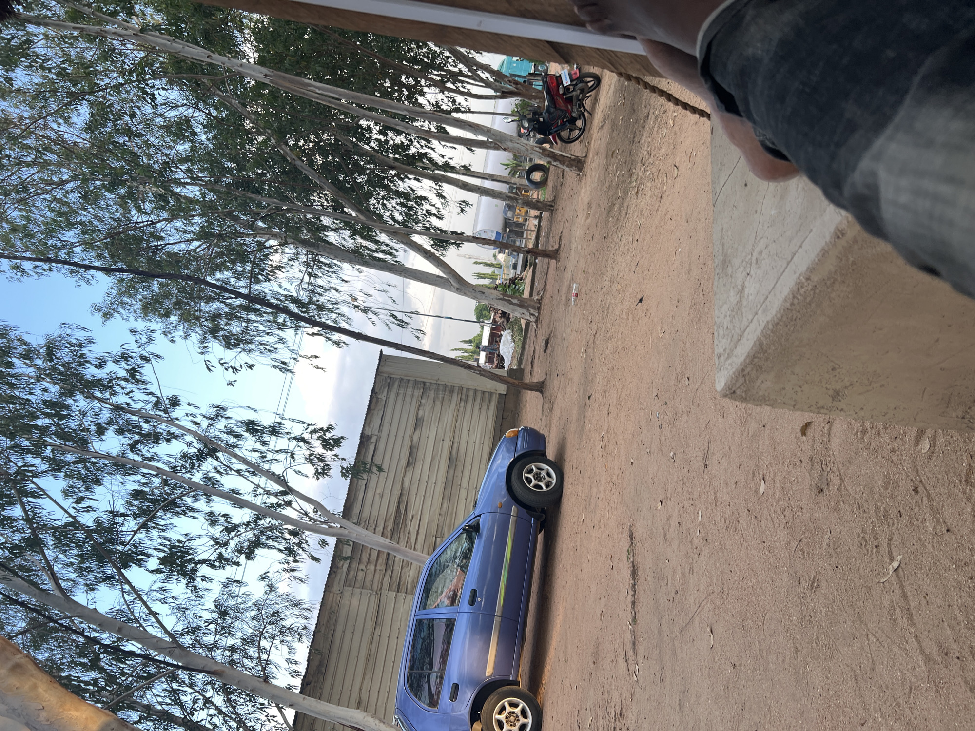 In Mayo Dasa, Nigeria, an outdoor scene unfolds with a clear sky and sparse trees. A small blue car is parked on a dirt lot, its side facing the viewer. Behind the car, a corrugated metal building stretches across the frame. In the distance, various vehicles are visible, including a white tanker truck, a red motorcycle, and other trucks. The foreground shows a person's legs and feet, clad in denim jeans, resting on a concrete structure. The ground is covered in dirt and scattered leaves, with a plastic bottle lying near the car. The overall impression is one of a rustic, functional area, possibly a roadside stop or a rural commercial space.