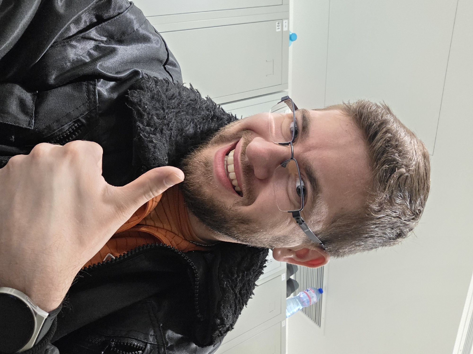 A smiling man with glasses and a beard is giving a thumbs-up. He is wearing a black jacket with a fuzzy collar and an orange shirt underneath. He has a smartwatch on his left wrist. The background appears to be an office setting, with filing cabinets visible behind him. There is a water bottle sitting behind him. The location is likely not in Plovdiv, Bulgaria, as there are no indicators to confirm this.