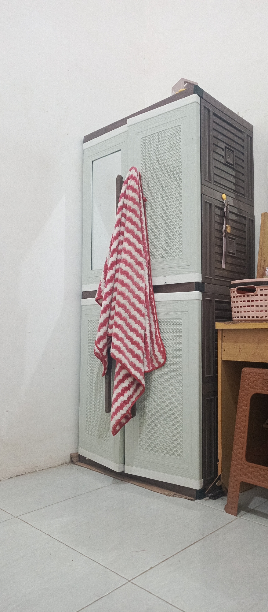 A vertically oriented, eye-level medium shot captures a corner of a sparsely furnished room. Dominating the center is a two-tone plastic wardrobe, light green on the left and dark brown on the right. A red and white striped towel is draped over the left half of the wardrobe, hanging from a hook. The wardrobe stands on a light-colored tiled floor. To the right of the wardrobe, a small wooden table with a yellow surface holds a pink woven basket. A portion of a brown plastic stool is visible in the lower right corner. The wall behind the wardrobe is plain white. The lighting is soft and diffused, suggesting ambient light. No people are present.