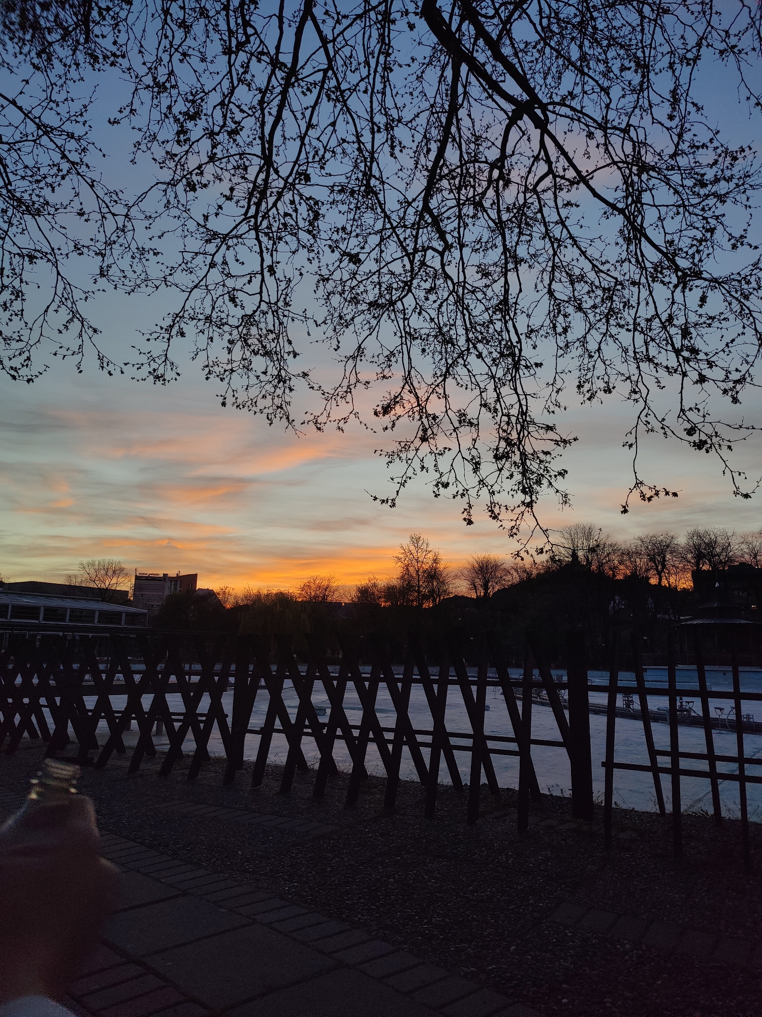 The image is taken at dusk or dawn, with a view overlooking a fence toward a body of water, likely the Rowing Canal in Plovdiv, Bulgaria. The sky features a gradient of colors, transitioning from blue at the top to orange and pink hues on the horizon. Bare tree branches silhouette against the sky. A dark picket fence runs horizontally across the midground, with the body of water visible behind it. The Rowing Canal has patches of ice on it. The foreground is dark and out of focus. Buildings and trees are visible on the horizon. A hand with a bottle is in the lower left corner of the image.