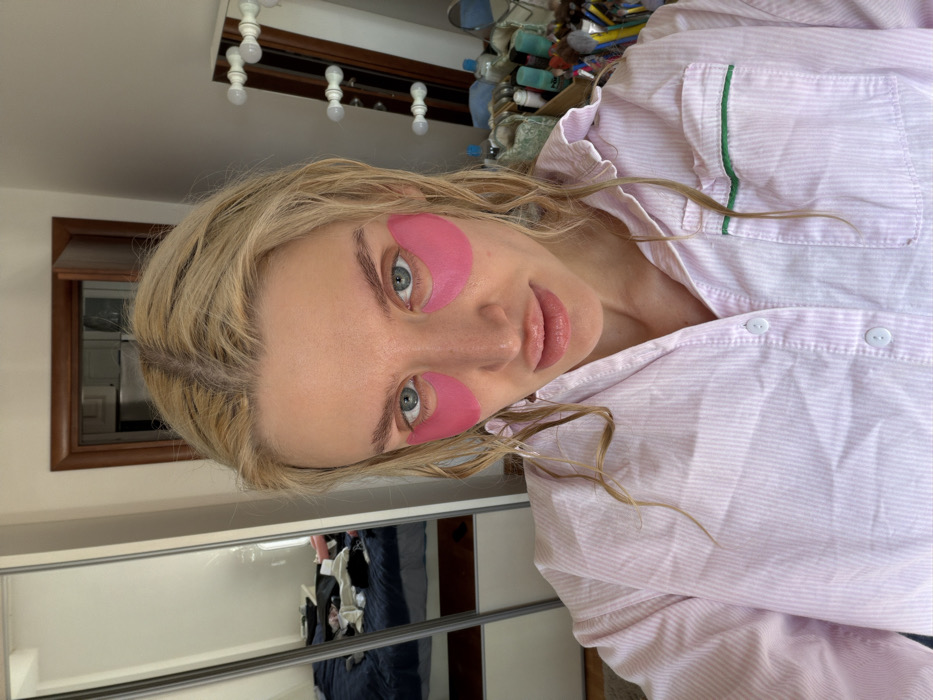 An indoor, close-up photograph depicts a female subject from the chest up, centered in the frame. The subject has blonde, wet-appearing hair parted in the middle and blue eyes. Two pink, crescent-shaped under-eye patches are applied beneath her eyes. Her lips appear full with a glossy finish. She is wearing a light pink, vertically striped button-up shirt featuring a single left chest pocket with a green trim. The subject looks directly into the camera with a neutral expression.
The background elements are visible behind the subject. To the left, a large, two-panel sliding mirror wardrobe reflects a messy room, including a bed with a dark blue comforter and various articles of clothing piled on it. Above the bed reflection, a light-colored wall is visible. In the upper center, a dark wooden door frame is partially visible, with a reflection within the doorway suggesting another room or a metallic appliance. To the right, a white vanity mirror with multiple exposed spherical light bulbs along its frame is present. Below the vanity mirror, a cluttered surface holds numerous bottles, jars, and makeup brushes, indicative of a cosmetic or skincare collection. A small section of a light-colored floor or rug is visible at the bottom left. The exact location within Budva, Montenegro, is not discernible from the visual information provided in the image.
