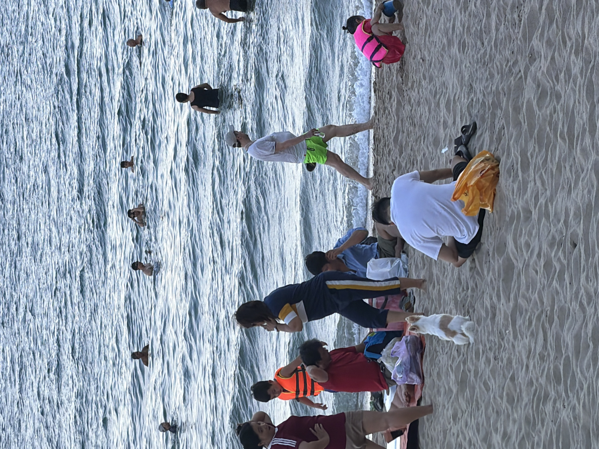 The image depicts a crowded beach scene in Nha Trang Ward, Vietnam. Several people are in the ocean, with some partially submerged and others swimming. On the shore, individuals are seen sitting, standing, and interacting. Some are dressed in swimwear, while others wear casual clothing. Objects present include beach towels, bags, and a life jacket worn by a child. The environment is characterized by sand and the sea.
