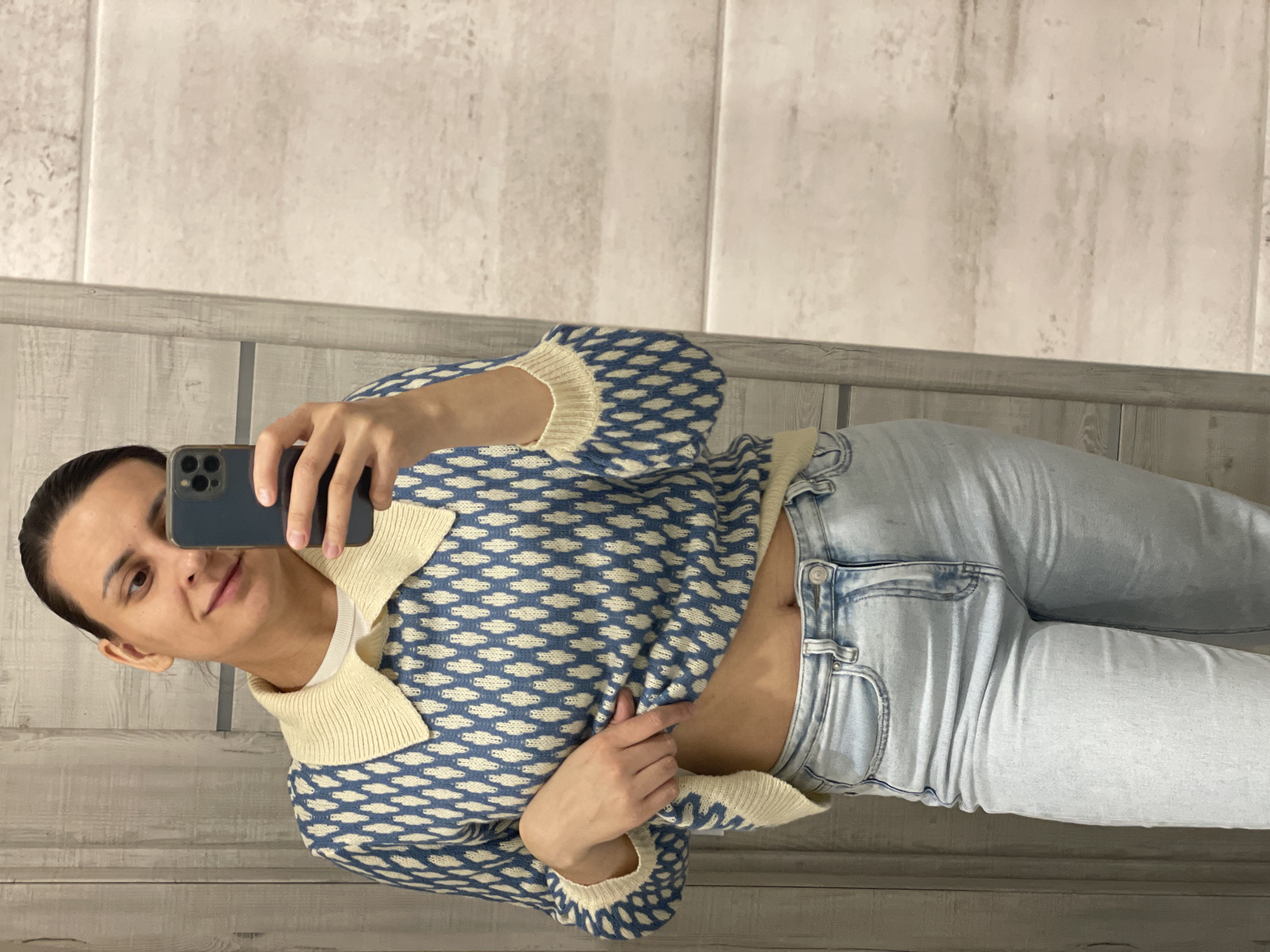A woman is taking a selfie in front of a bathroom mirror. She is wearing a blue and white patterned sweater with a beige collar and light wash jeans. Her hair is pulled back, and she is holding a dark-colored smartphone in her right hand. The bathroom walls are decorated with light-colored, textured tiles, and there is a gray wooden trim at the bottom. The location is Unknown, Unknown.
