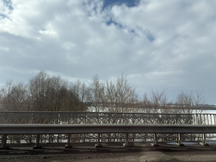 The image displays an elevated outdoor scene, identified as Yus'ki, Russia. The upper half of the frame is filled with a predominantly cloudy sky, featuring a mixture of grey-white clouds and interspersed patches of clear blue. A prominent grey metal guardrail, secured by dark bases, runs horizontally across the lower third of the image. Immediately behind and partially obscured by the guardrail, a decorative metal railing with an intricate, repetitive scrollwork pattern extends across the frame. Below these railings, the ground surface appears dark and uneven. Beyond the railings, a dense line of bare, dormant deciduous trees occupies the midground, suggesting a cold season. The background reveals a vast, flat expanse uniformly covered in white snow, stretching towards a low, indistinct horizon. No individuals, vehicles, or active elements are present within the static scene, which is illuminated by diffuse daylight.
