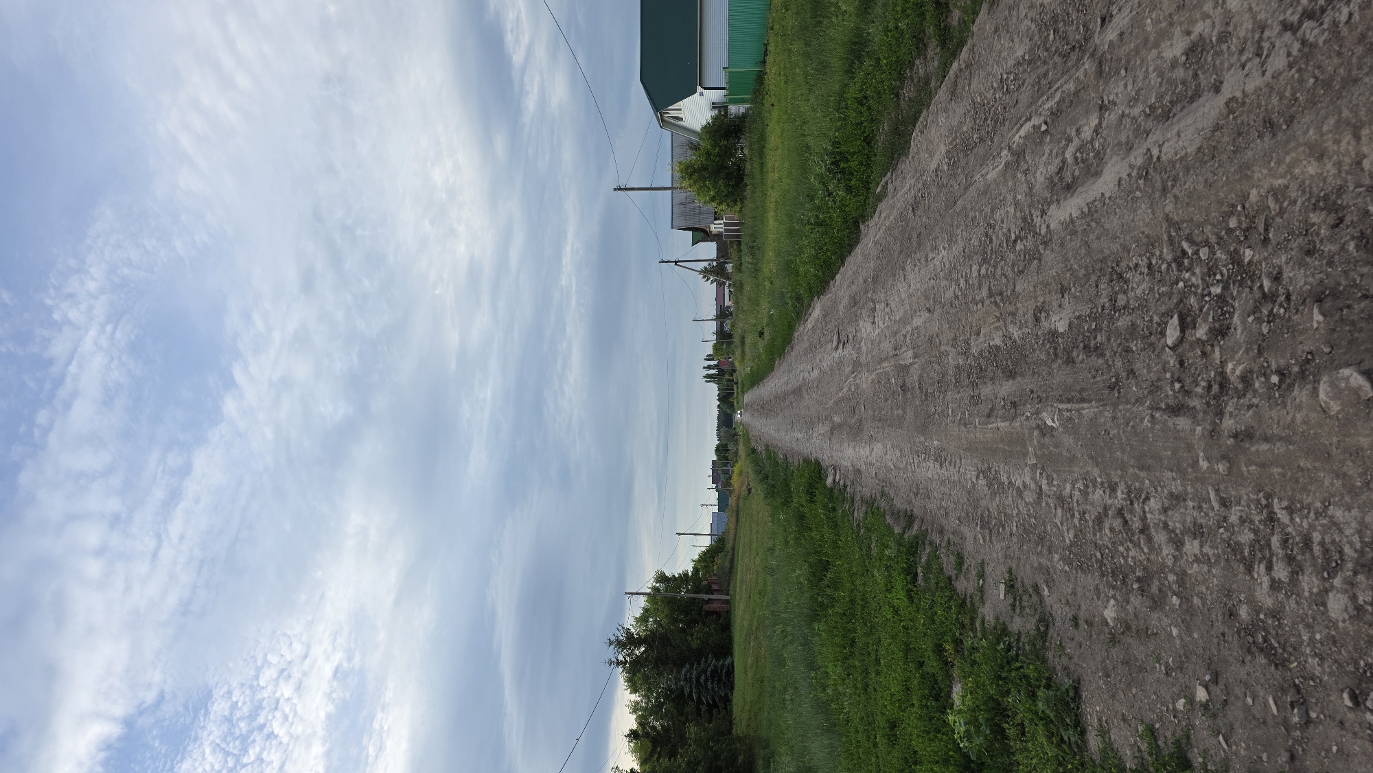 The image depicts a rural dirt road in the village of Kupoyarovo, Russia, leading into the distance under a cloudy sky. The road is unpaved and shows signs of wear, with visible tire tracks and scattered stones. Lush green grass lines both sides of the road. Several houses with gabled roofs and green fences are visible along the right side of the road, interspersed with trees and utility poles. A few trees are also present on the left side of the road, with more houses and structures further in the distance. The sky is filled with a mix of light and dark gray clouds. A white vehicle is faintly visible on the road in the far distance.