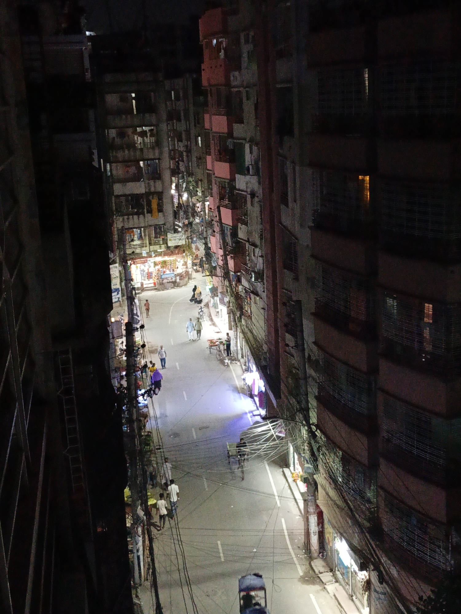 A high-angle, nighttime view down a street in Dhaka, Bangladesh. The street is lined with tall buildings on both sides, some with balconies. Many people are walking and some are standing on the street. In the upper portion of the view, a store is well lit. Some power lines and electrical wires are visible on the side of the street. There is at least one rickshaw on the road.