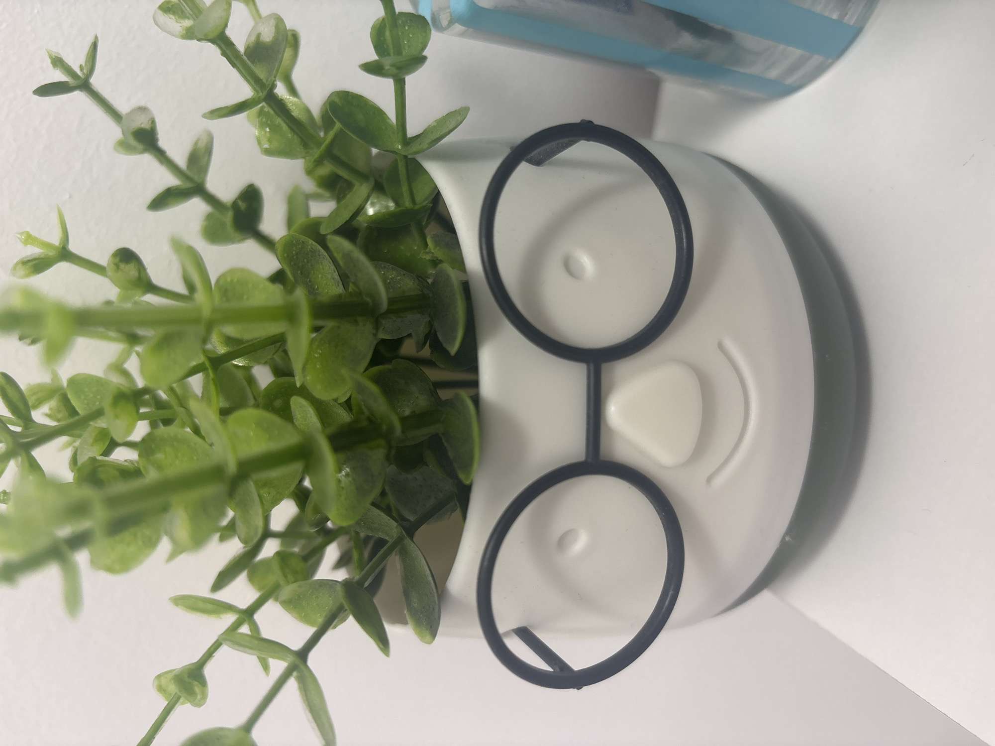 The image presents a close-up, eye-level view of an artificial plant contained within a white ceramic planter, designed to resemble a stylized face. The planter features two circular black wire-rimmed glasses, a raised triangular nose, two subtle circular indentations functioning as eyes, and a carved, upward-curving smile. Its base is a darker gray-green hue.
The plant itself consists of multiple thin green stems adorned with numerous small, rounded, light green leaves. Many leaves display a slight glossy texture and appear to have minuscule water droplets or reflections on their surface.
The planter rests on a flat, white, and slightly reflective surface, likely a shelf or desk. Behind the planter is a plain white wall. To the right, partially visible, is a vertical object with alternating light blue and white stripes, the specific nature of which cannot be fully determined from the crop.
The scene is brightly and evenly lit, characteristic of an indoor setting. The foreground and main subject are in sharp focus, while the upper portions of the plant and the striped object on the right exhibit a slight blur. No identifiable markers or features within the image indicate a specific location in the city of Unknown, Unknown.