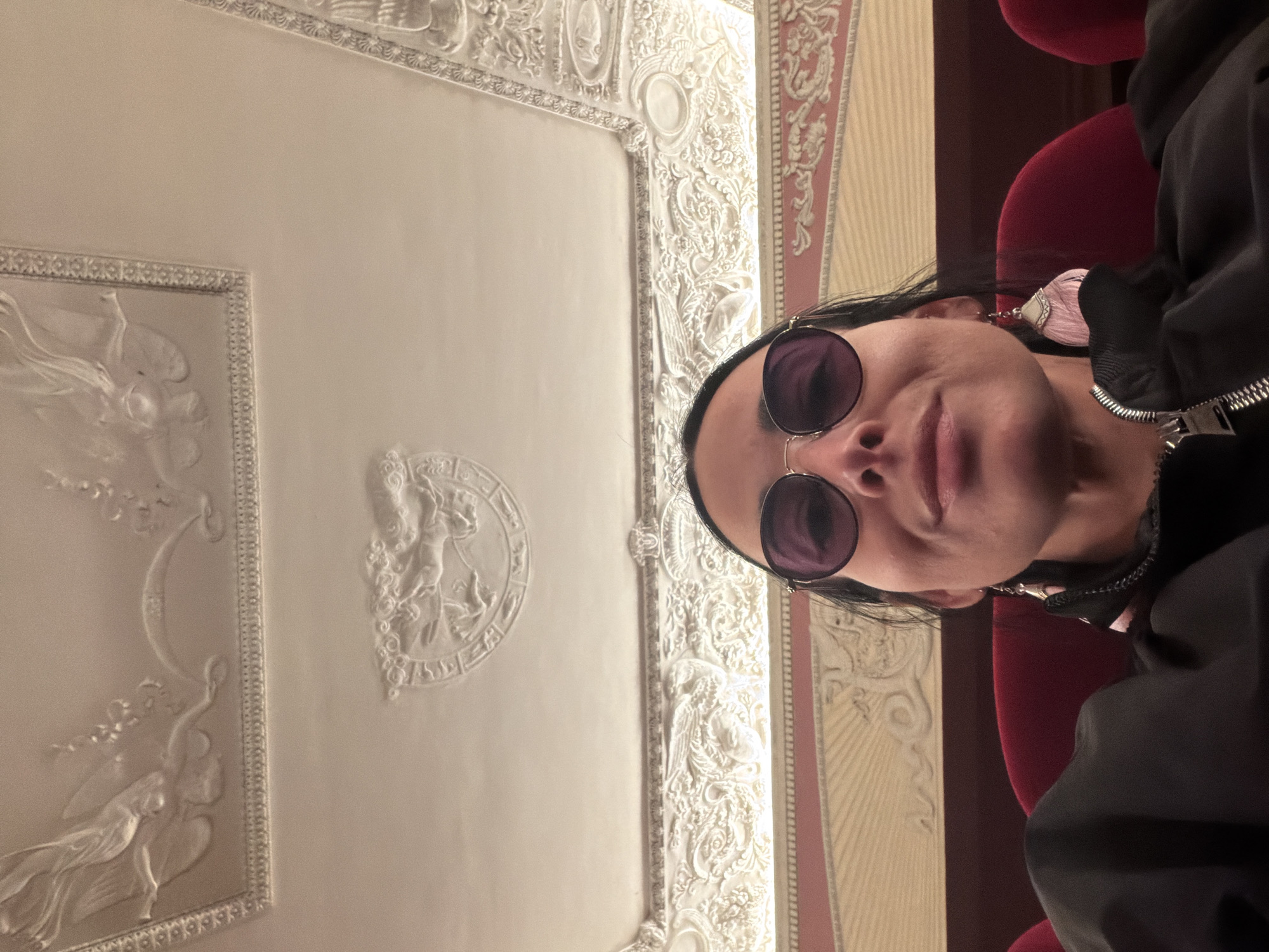 The image captures a vertical selfie of a person looking upwards in an ornate interior, set in Saint Petersburg, Russia. The subject is a person, likely female, with dark hair, wearing dark, round sunglasses with gold-colored frames, and a dark jacket with a silver zipper. A prominent, elaborate pink earring with a tassel-like or textured design hangs from their left ear. Their lips are slightly parted, suggesting a subtle smile or relaxed expression.
The dominant background feature is a highly decorated white ceiling, showcasing intricate stucco or plaster work. It includes rectangular panels depicting classical figures, possibly angels or cherubs, with flowing drapery, and a central circular medallion also featuring a figure. The detailed craftsmanship points to a grand architectural style, such as Baroque or Neoclassical.
To the right of the person, a section of a wall or column is visible, featuring a patterned design in reddish-pink and cream tones, possibly with vertical stripes and classical motifs. Behind the person's shoulder, a plush red velvet seat or part of a chair can be seen, suggesting a formal or theatrical setting. The overall scene indicates an opulent interior, likely within a historic building like a theater, opera house, or palace. The lighting is soft and even, typical of indoor artificial illumination. No visible text is present in the image.