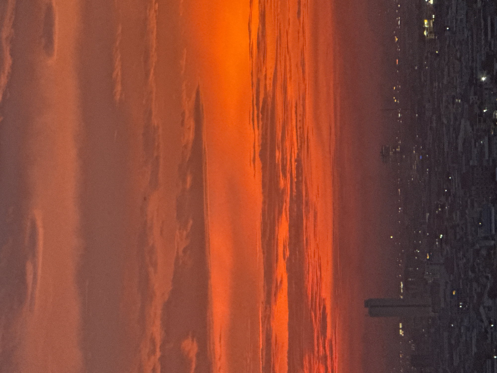 The image captures a sunset over the cityscape of Khan Mean Chey, Cambodia. The sky is dominated by vibrant hues of orange and red, with distinct bands of clouds creating a textured effect. Below the fiery sky, the city is depicted in silhouette, with buildings and lights faintly visible. A prominent, tall building stands out in the mid-ground, appearing as a dark rectangular shape against the twilight sky, with illuminated windows. Numerous smaller lights dot the urban landscape, indicating a densely populated area. The overall scene is one of an urban environment transitioning into night under a dramatic, colorful sky.