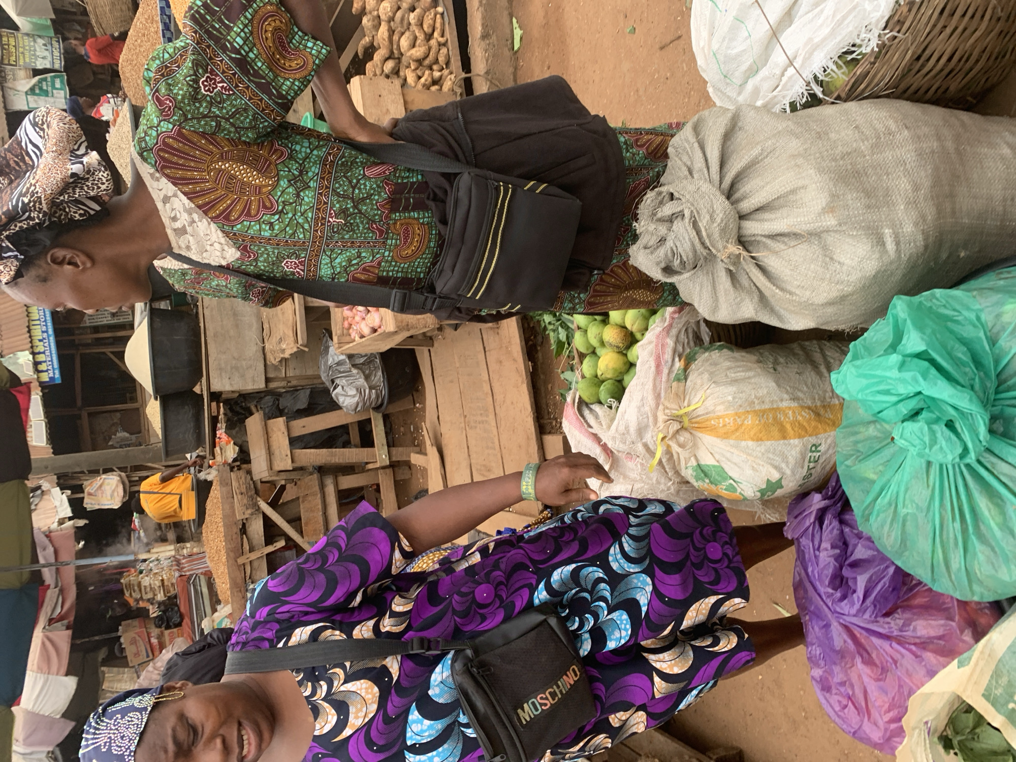 A medium shot captures two Nigerian women at a vibrant outdoor market stall in Shagamu, Nigeria. The woman on the left, with a purple and blue patterned headscarf and a matching patterned dress, is visible from the chest up, her face contorted in a grimace, her mouth slightly open. She wears a black cross-body bag with "MOSCHINO" printed on it. The woman on the right, wearing a green and brown patterned dress and a leopard-print headscarf, is seen from the chest down, her body turned slightly away from the camera. She has a black bag with gold stripes slung across her shoulder.
Surrounding the women are various market goods. In the foreground, to the left, are several colorful plastic bags: one purple, one bright green, and a partial view of a white bag containing green leaves. Behind these, to the right, are sacks of produce, including a large white sack with yellow banding and text that appears to read "MASTER", and a large, rough-textured grey sack tied at the top. A woven basket filled with green produce is partially visible to the far right. In the middle ground, visible between the two women, are several bunches of green fruits, likely limes or small mangoes, and more sacks of grains or dried goods. The background reveals the bustling market environment with wooden stalls, awnings, and other market-goers in the distance, including a person in a yellow shirt tending to a large pile of what appears to be dried shrimp or small fish. The ground is dirt.