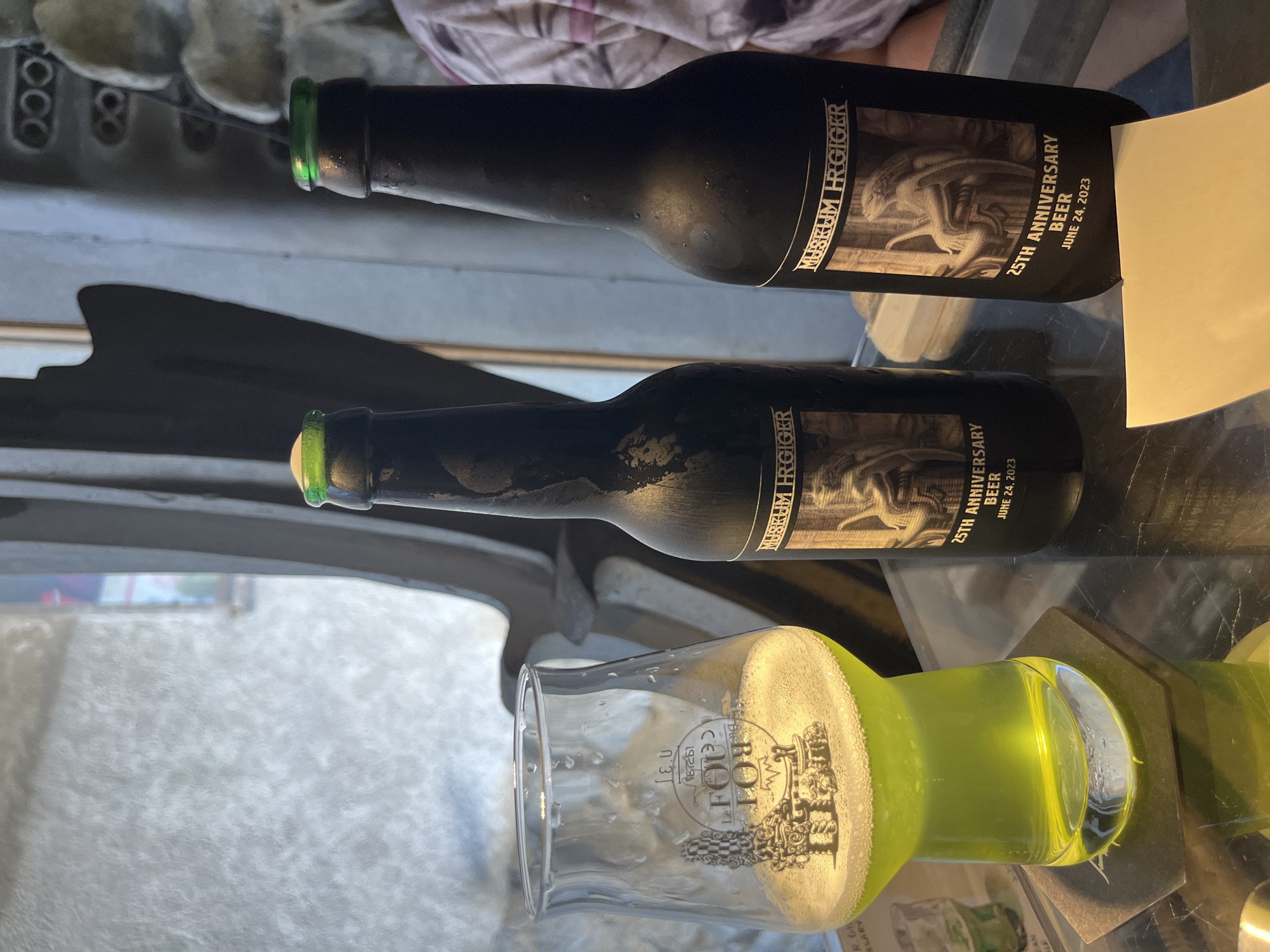 Two bottles of "MUSEUM HR GIGER 25TH ANNIVERSARY BEER JUNE 24, 2023" are prominently displayed on a glass surface, one slightly in front of the other. The bottles are dark with frosted glass and have labels featuring an illustration of a seated figure. A glass of pale green liquid, likely beer, sits to the left, partially filled, with a frothy head. The glass has a logo that appears to read "LE FOULOY" with an intricate illustration. The background is out of focus, showing a textured, light-colored wall or surface on the left and a dark, sculptural element on the right. A person's arm and shoulder, wearing a horizontally striped garment, are visible in the far right background. The scene appears to be indoors, possibly in a bar or museum setting in Epagny, Switzerland.