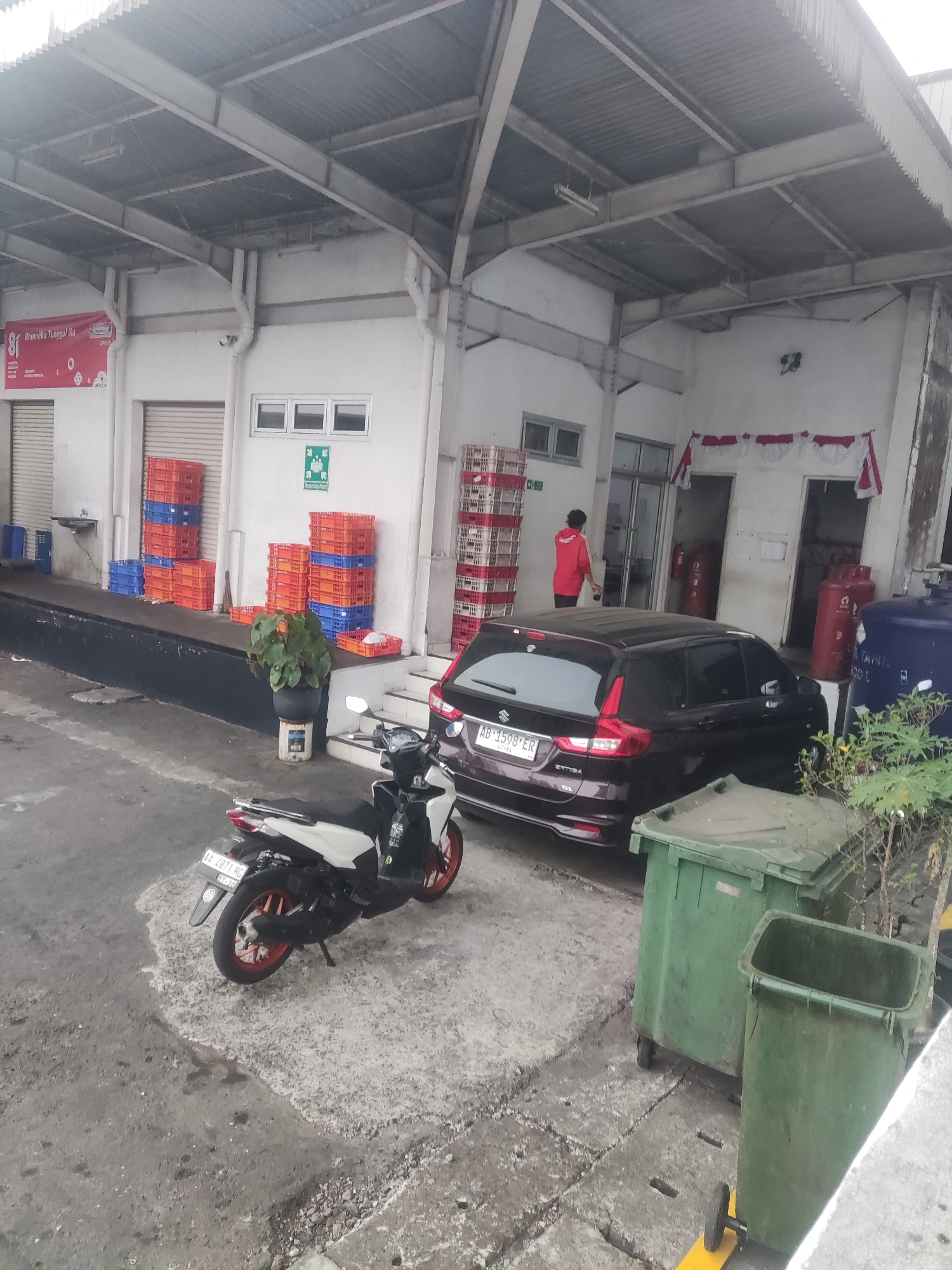 In an outdoor loading dock area, a dark brown Suzuki Ertiga is parked in front of a building with a corrugated metal roof supported by steel beams. A white and black motorcycle with orange accents is parked to the left of the car. In the foreground, two large green trash bins with wheels are visible. The building has a white facade with several windows and doors. Stacks of orange and blue plastic crates are piled near the entrance. A person wearing a red shirt is standing near an open doorway, and a large blue water tank is situated to the right of the building. The ground is paved with concrete and gravel. The scene appears to be in Trihanggo, Indonesia.