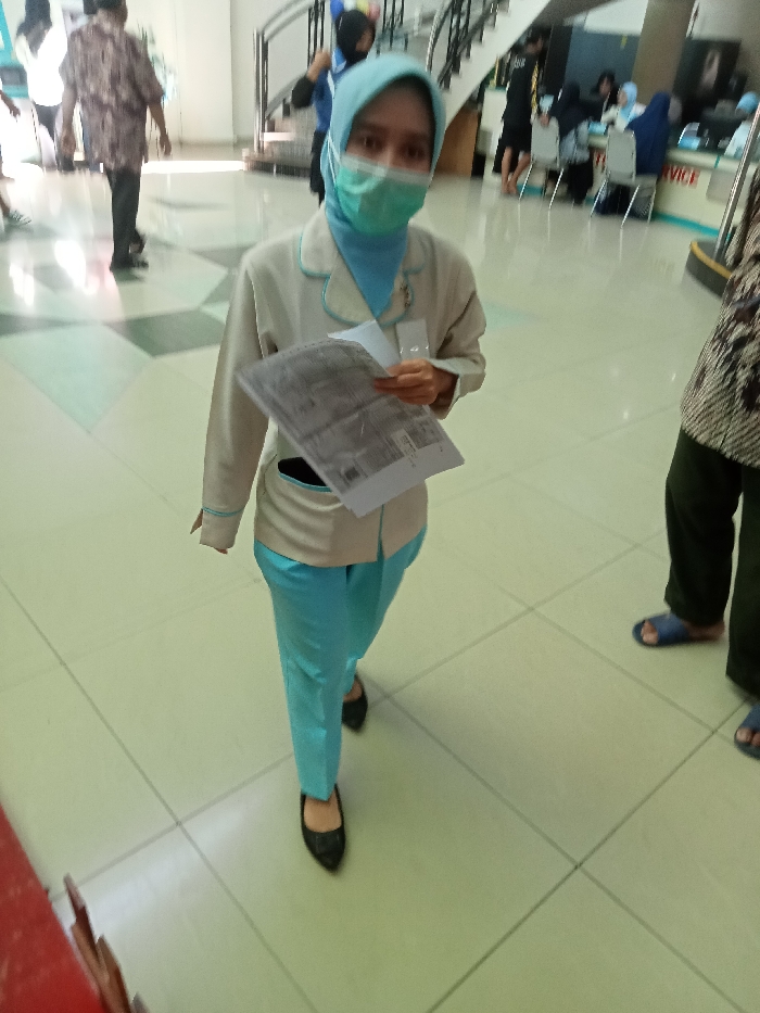 The image shows a young woman wearing a white jacket with a blue collar, a blue hijab, and a blue surgical mask. She is holding a white paper in her right hand, looking down at it. She is standing on a tiled floor inside what appears to be a public building. The floor is light green and beige, with white square tiles. To the right, there is another person wearing batik pants, and their feet are visible, one wearing a blue sandal. The location is likely a government building or office in Kecamatan Margorejo, Indonesia, based on the coordinates provided. The young woman appears to be standing in a waiting area, possibly at a counter or service desk, as there are other people visible in the background.
